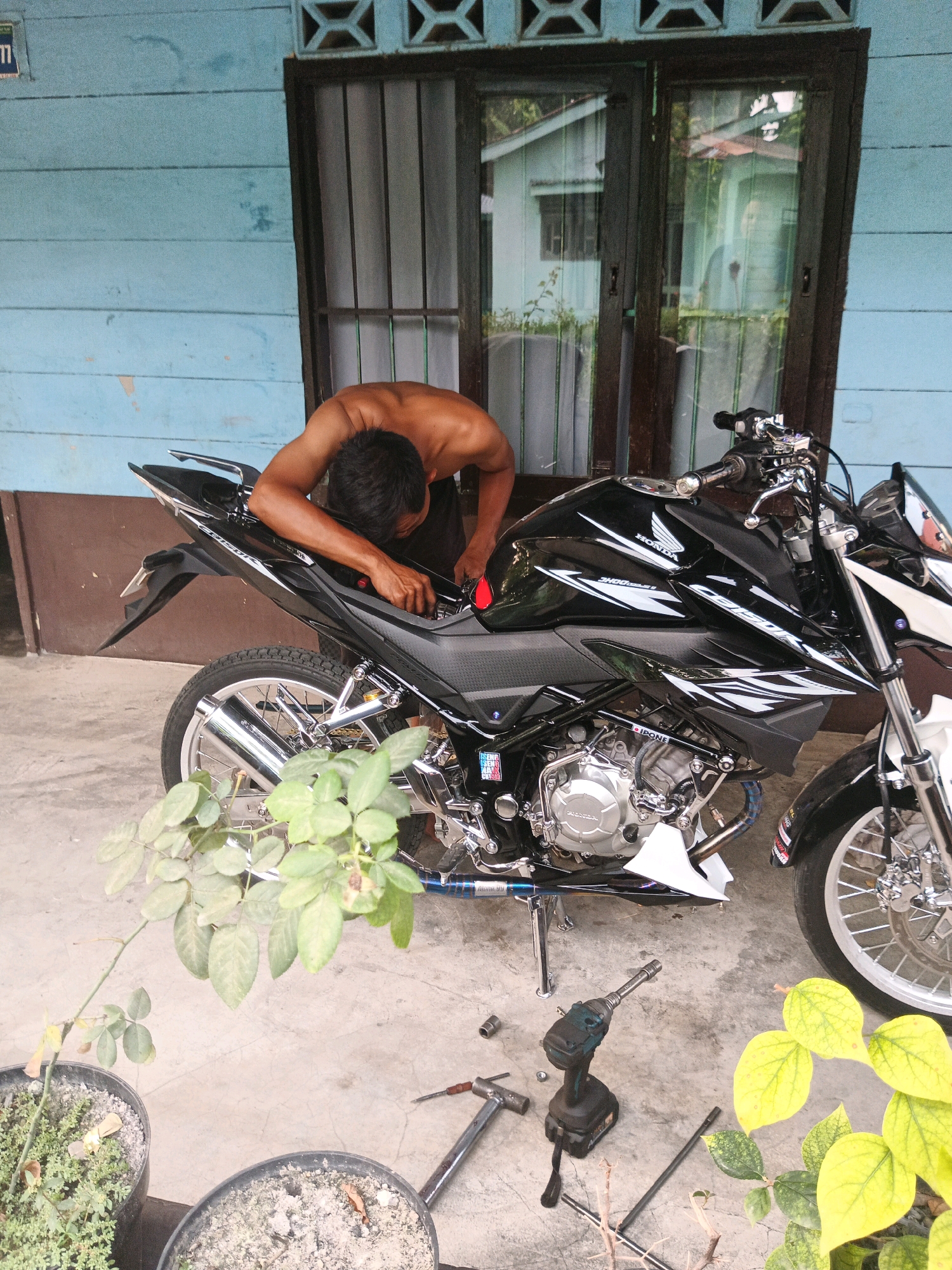 A shirtless man is seen working on a black motorcycle parked on a concrete floor in front of a light blue wooden building. The man is leaning over the rear of the motorcycle, with his head bent down and his hands engaged in the motorcycle's mechanics. Tools, including a drill and a wrench, are scattered on the ground near the motorcycle. Potted plants are present on the left and right sides of the scene. The building has a window with dark frames and vertical bars. The overall scene suggests an outdoor maintenance or repair activity. The location is identified as Linggatiga, Indonesia.