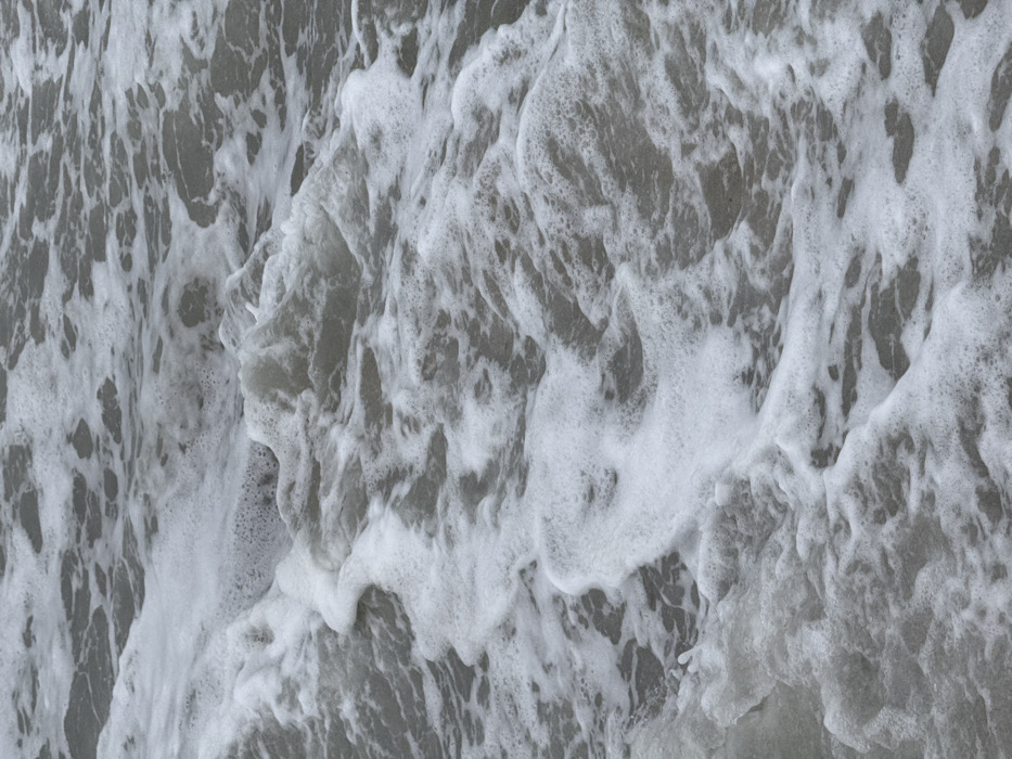 The image presents a close-up, top-down view of agitated water, likely a coastal area in Vila Nova de Gaia, Portugal. The scene is dominated by a grayscale palette, with dark grey water interspersed with numerous irregular patches and streaks of bright white sea foam. The foam varies in density, from frothy accumulations forming ridges and peaks to thinner, more dispersed bubbly layers. The pattern of the foam suggests ongoing wave action, with some areas appearing to be a result of breaking waves and others as receding wash. No individuals, animals, or man-made objects are visible within the frame. The immediate environment is solely the water surface.
