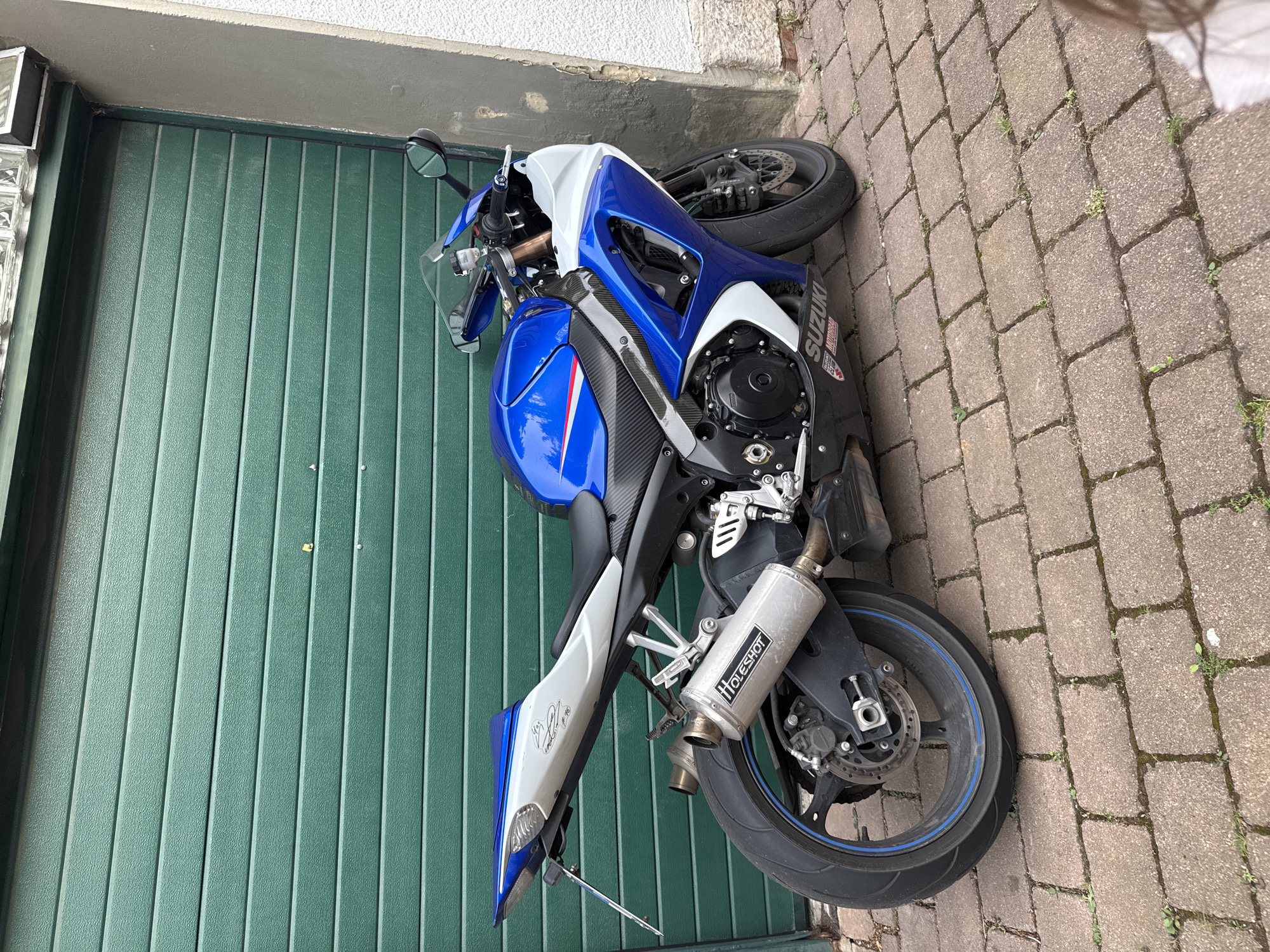 A blue and white Suzuki sport motorcycle is parked on a brick paver pathway in front of a closed, green, slatted garage door. The motorcycle is positioned at an angle, with its front facing towards the right of the image. The motorcycle features a blue fuel tank and fairings with white accents, and a silver tail section. The exhaust pipe on the right side is visible and has the "HoleShot" logo on it. The garage door is made of horizontal metal slats and appears to be the main backdrop. To the right of the garage door, a portion of a rough, grey-colored wall is visible. The ground is covered in reddish-brown brick pavers, with some small patches of green moss and weeds growing in the cracks. The lighting suggests it is daytime. No people are clearly visible in the frame, although a small portion of a person's head and shoulder, possibly a child, is visible in the lower right corner, out of focus.