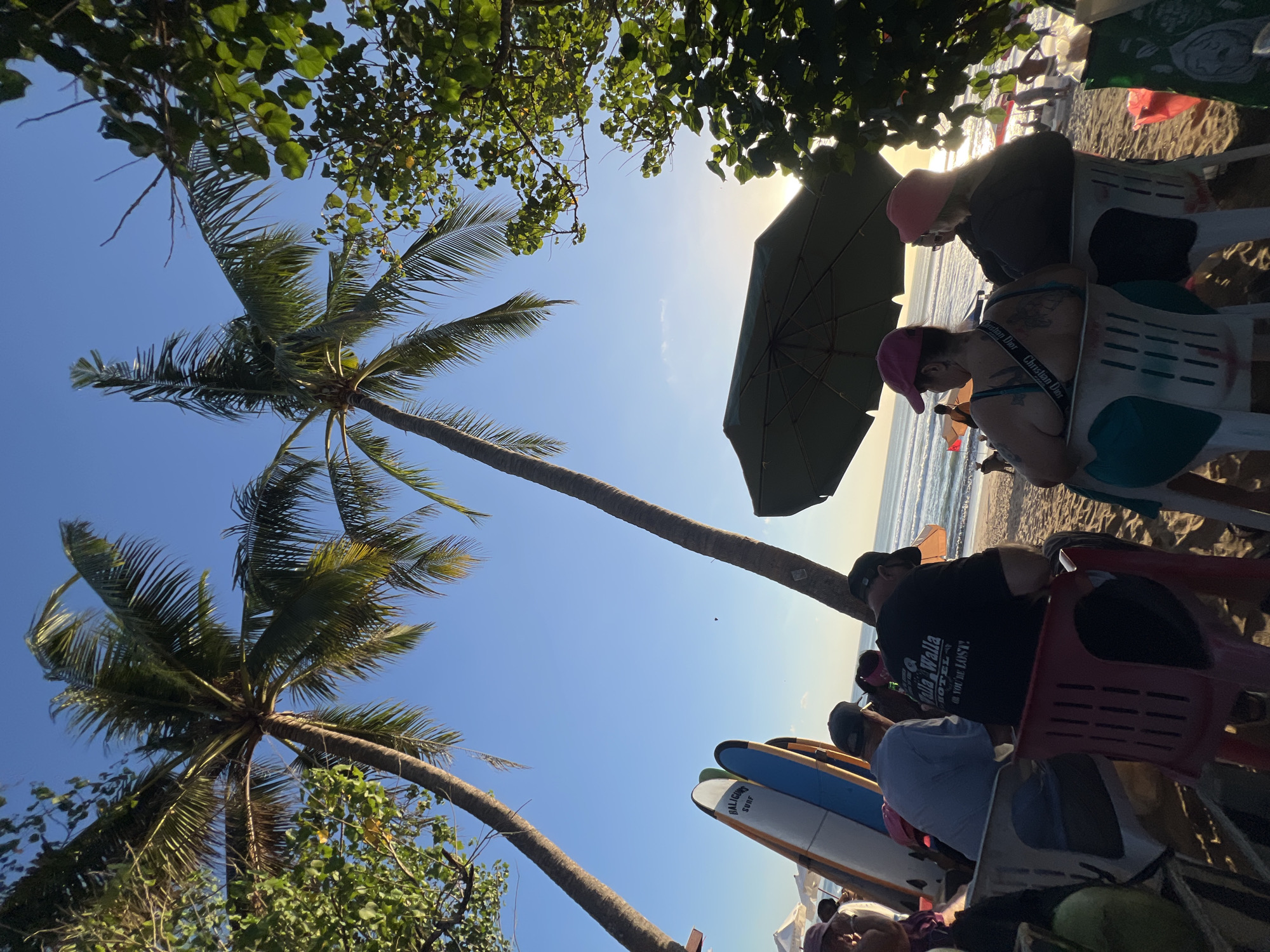 In Kuta, Indonesia, a beach scene unfolds under a clear blue sky, with the sun setting and casting a warm glow. Two tall palm trees frame the view, their fronds swaying gently. In the foreground, several people are seated on chairs on the sandy beach, facing the ocean. One man wears a black t-shirt with white lettering that reads "Walla Walla Hotel - You're Lost!". Next to him, another person in a light blue shirt and a cap is also seated. In front of them, two surfboards stand upright. Further back, more people are visible enjoying the beach, with umbrellas and beach chairs dotting the sand. The ocean stretches to the horizon, with gentle waves rolling in. The overall atmosphere is relaxed and peaceful, with a hint of tropical charm.
