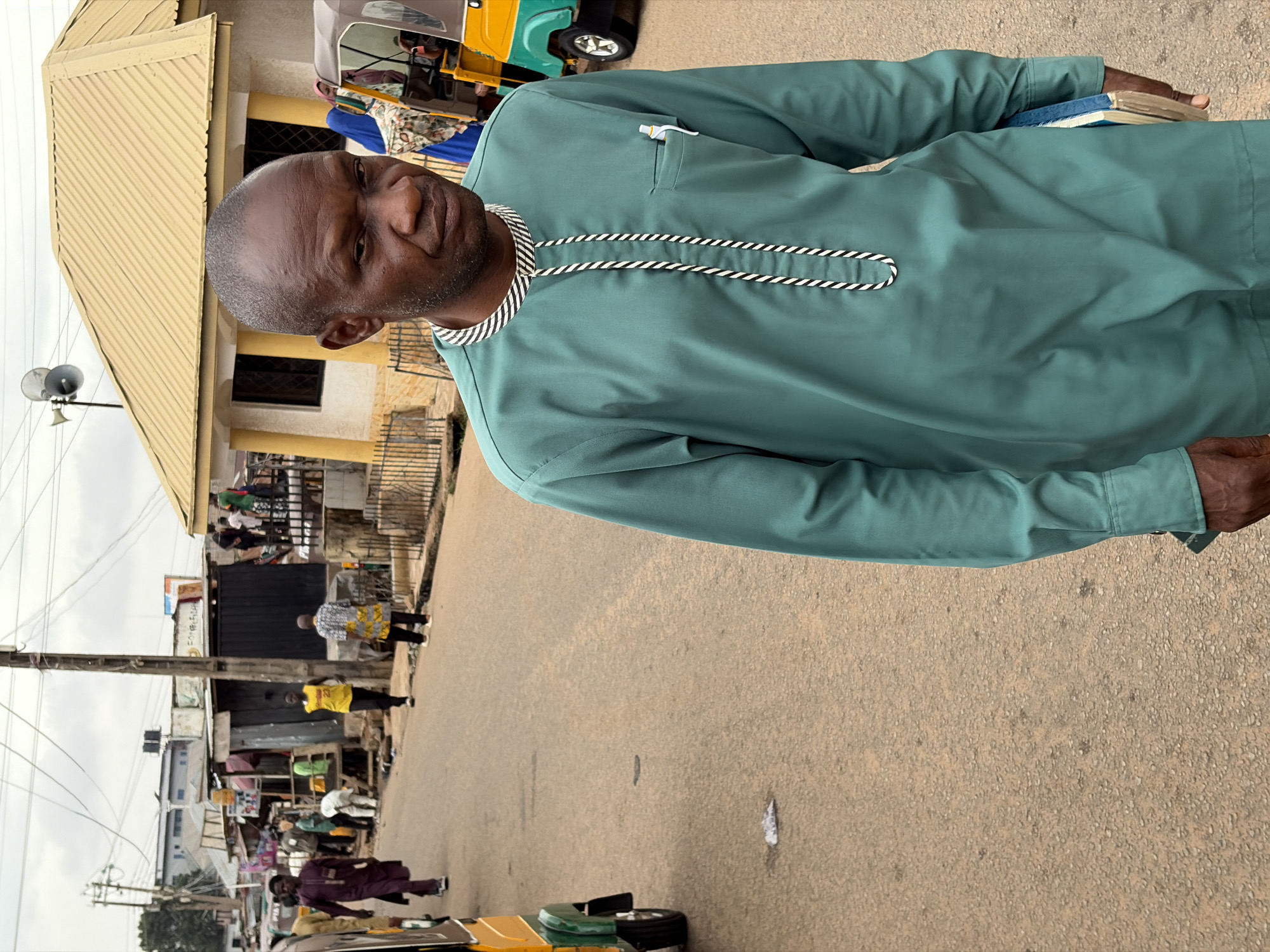 A man wearing a green long-sleeved shirt with black and white rope trim on the collar and chest stands in a street in Jos, Nigeria. He is holding a small stack of books in his left hand. Behind him, there is a building with a light-colored roof and walls. A loudspeaker is attached to a pole in front of the building. Several people are visible in the background on the sidewalk and street. A tricycle taxi is parked nearby. The street is paved and appears somewhat worn. Power lines run overhead.