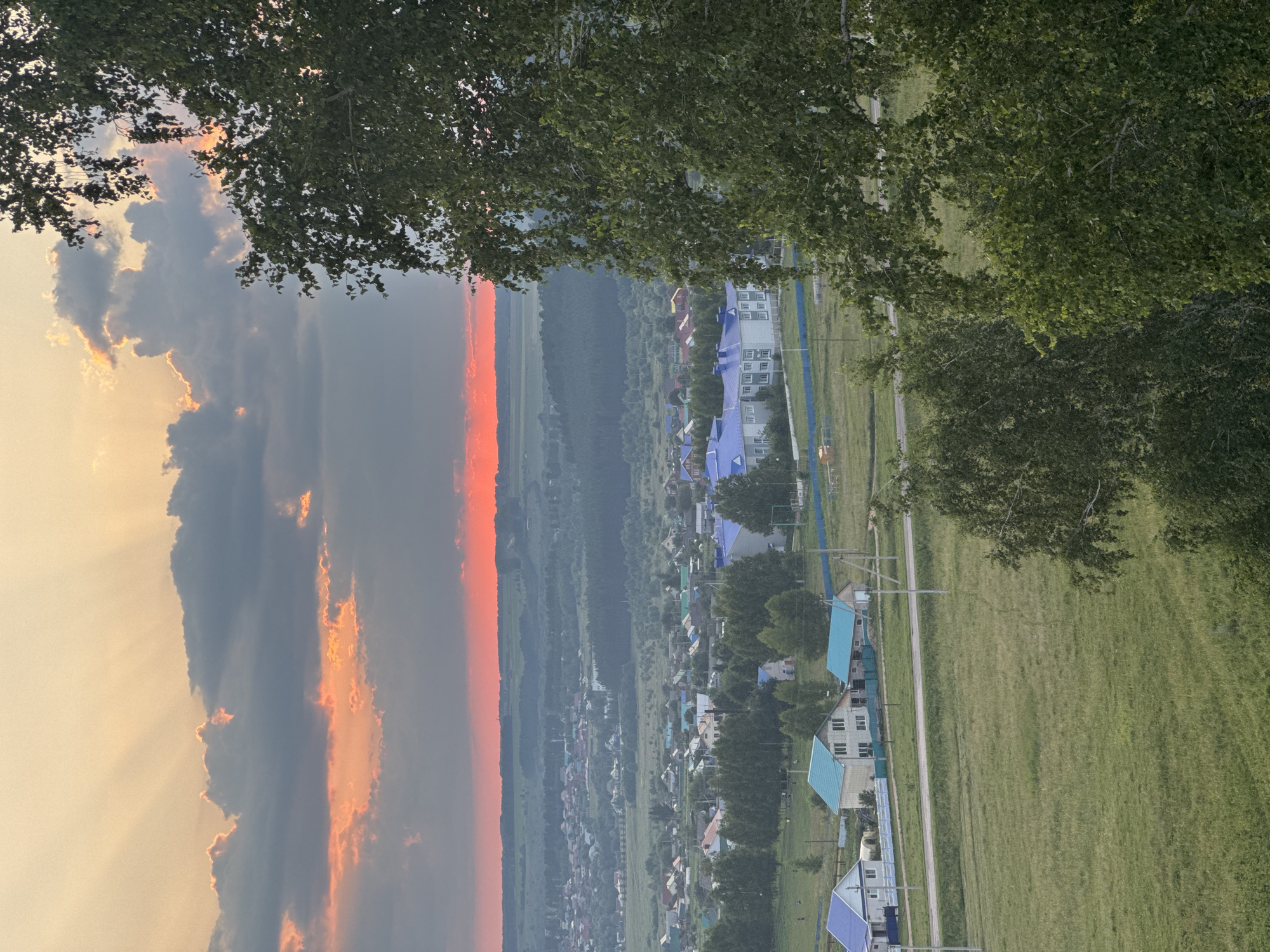 This is an aerial, wide-angle, vertical photograph taken at sunset in Sharan, Russia. The image depicts a rural landscape under a dramatic sky. The top portion of the frame is dominated by a sunset with vibrant orange and pink hues, partially obscured by dark, heavy clouds. Rays of sunlight pierce through the clouds, creating a crepuscular effect. A large, leafy green tree occupies the right side of the frame, its branches extending into the upper part of the image.
Below the sky, a sprawling village or town is visible. Numerous houses with various colored roofs, predominantly blue and teal, are scattered across the landscape. A few larger buildings, possibly institutional, stand out with their blue roofs. Fields of green grass and scattered trees intersperse the residential areas. Roads or pathways are visible, with utility poles lining some of them. In the distance, a hazy horizon suggests rolling hills or a flat, expansive plain. The overall scene conveys a sense of tranquility and the natural beauty of a rural sunset.