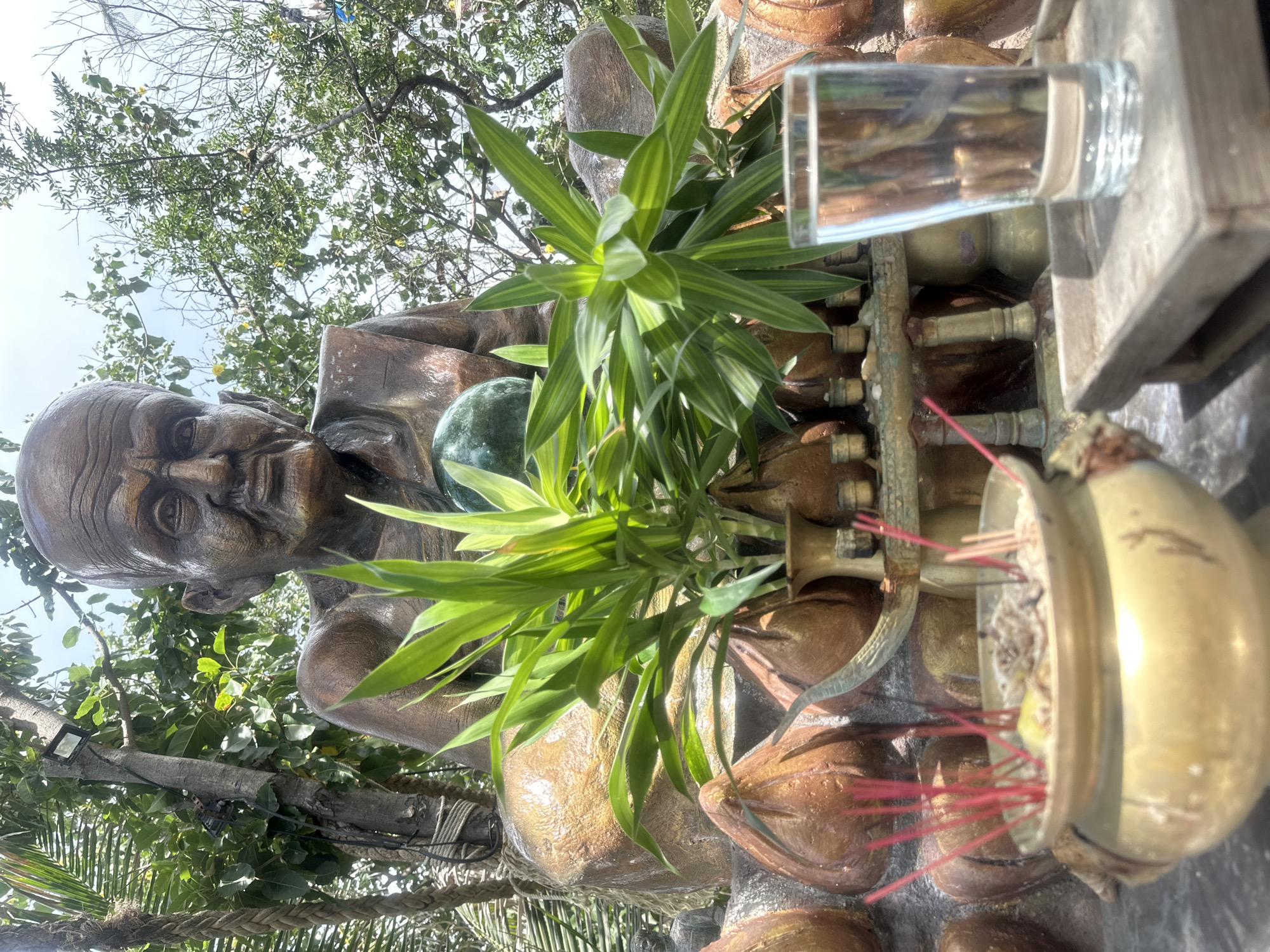 This is a medium shot, eye-level, outdoor photograph of a bronze statue of an elderly Buddhist monk, seated cross-legged with his hands resting on his lap. The statue is adorned with a green orb in its lap, and is surrounded by lush green foliage and trees. In the foreground, an incense pot with burning red incense sticks is visible, along with a glass of water placed on a wooden tray. The background is filled with trees and a bright, overcast sky. The scene appears to be in a temple or shrine in Ko Phangan, Thailand, a popular tourist destination known for its beaches and spiritual sites.