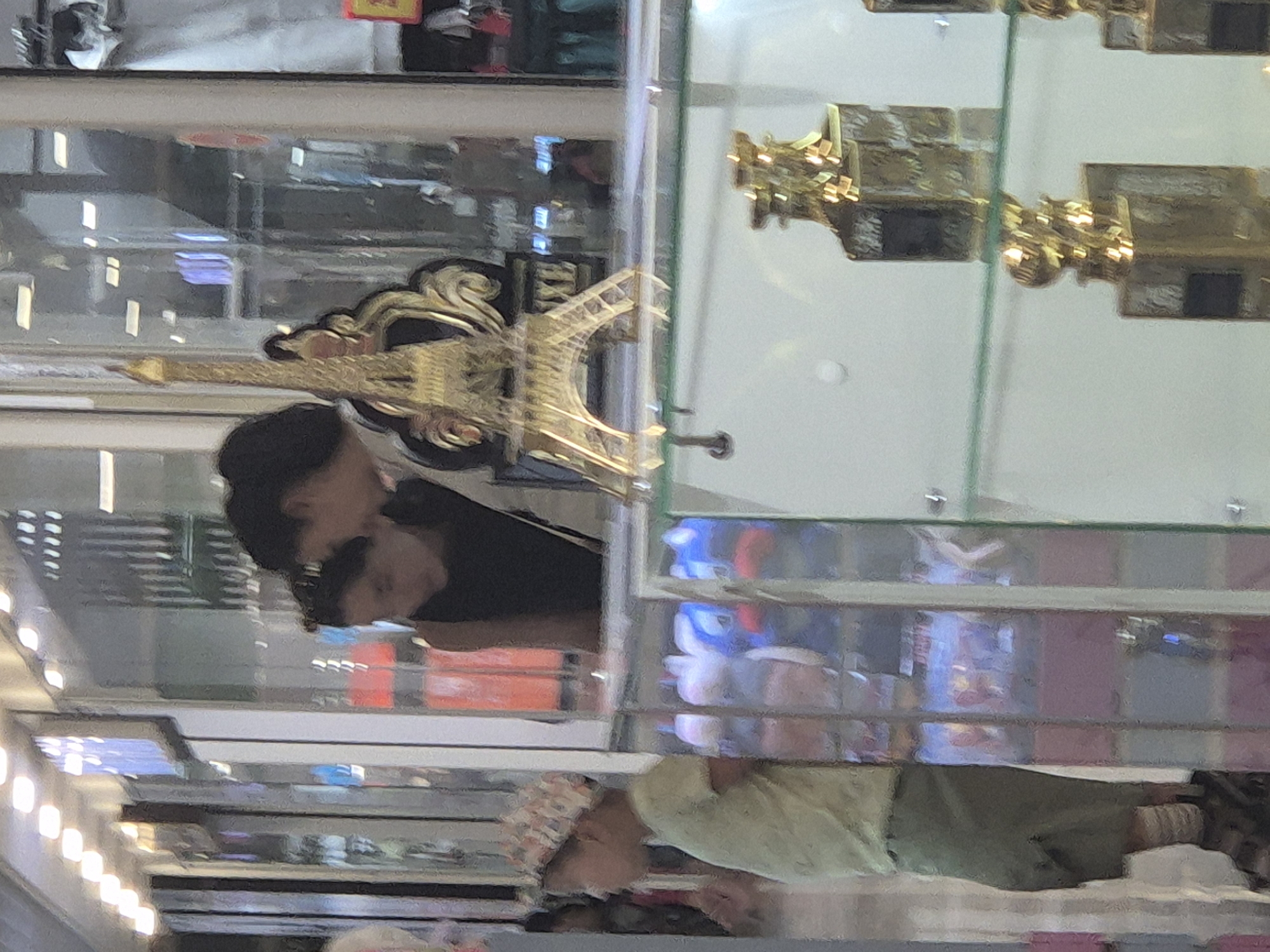 An indoor scene, possibly a market or shopping mall, shows a young child on the left side of the frame, wearing a light-colored t-shirt and a patterned cap. The child is looking to their left. To the right of the child, two adults are partially visible, looking down and to their left. In the foreground, a glass display case contains several gold-colored decorative items. One prominent item is a miniature replica of the Eiffel Tower. The background shows reflections of lights and displays, suggesting an indoor commercial environment. The image does not contain enough information to determine its location in Kokshetau, Kazakhstan.