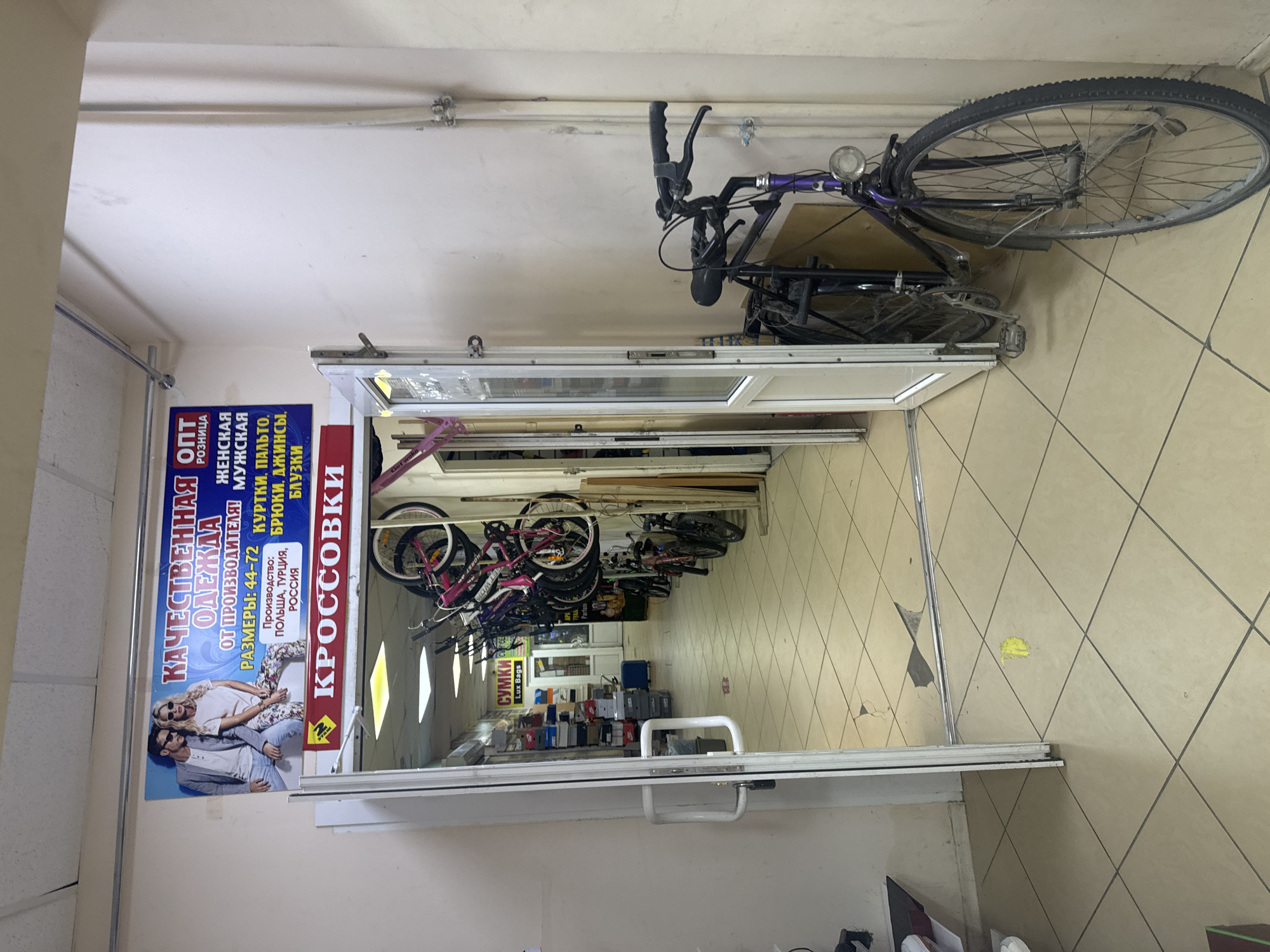 This image depicts the interior of a retail store, likely in Saint Petersburg, Russia, that sells clothing and footwear. The store appears to be a budget-friendly establishment, possibly an outlet or discount store, given the presence of signage advertising "OPT" (wholesale) and "ROZNITSA" (retail), as well as "OT PROIZVODITELYA" (from the manufacturer).
The main subjects visible are several bicycles, some of which are hanging from the ceiling or leaning against a wall. These appear to be in a state of disrepair or are simply being stored. In the foreground, there is a large advertisement for "KACHESTVENNAYA ODEZHDA" (Quality Clothing), featuring a man and a woman in casual attire. This sign also lists clothing types like "KURTKI, PALTO" (jackets, coats), "BRYUKI, DZHINSY" (trousers, jeans), and "BLUZKI" (blouses), along with production origins such as Poland, Turkey, and Russia, and sizes ranging from 44-72. Another prominent sign advertises "KOSSOVKI" (sneakers/trainers) and "SUMKI" (bags), with "Lux Bags" also visible.
The store's interior is characterized by tiled floors and a fluorescent-lit ceiling. Shelves stocked with boxes are visible in the background, suggesting merchandise storage or display. The overall impression is one of a functional, perhaps slightly cluttered, retail space focused on offering a variety of affordable goods. The lighting suggests it is daytime, and the setting is indoors. No specific events are depicted, but the scene captures the typical environment of a shop.