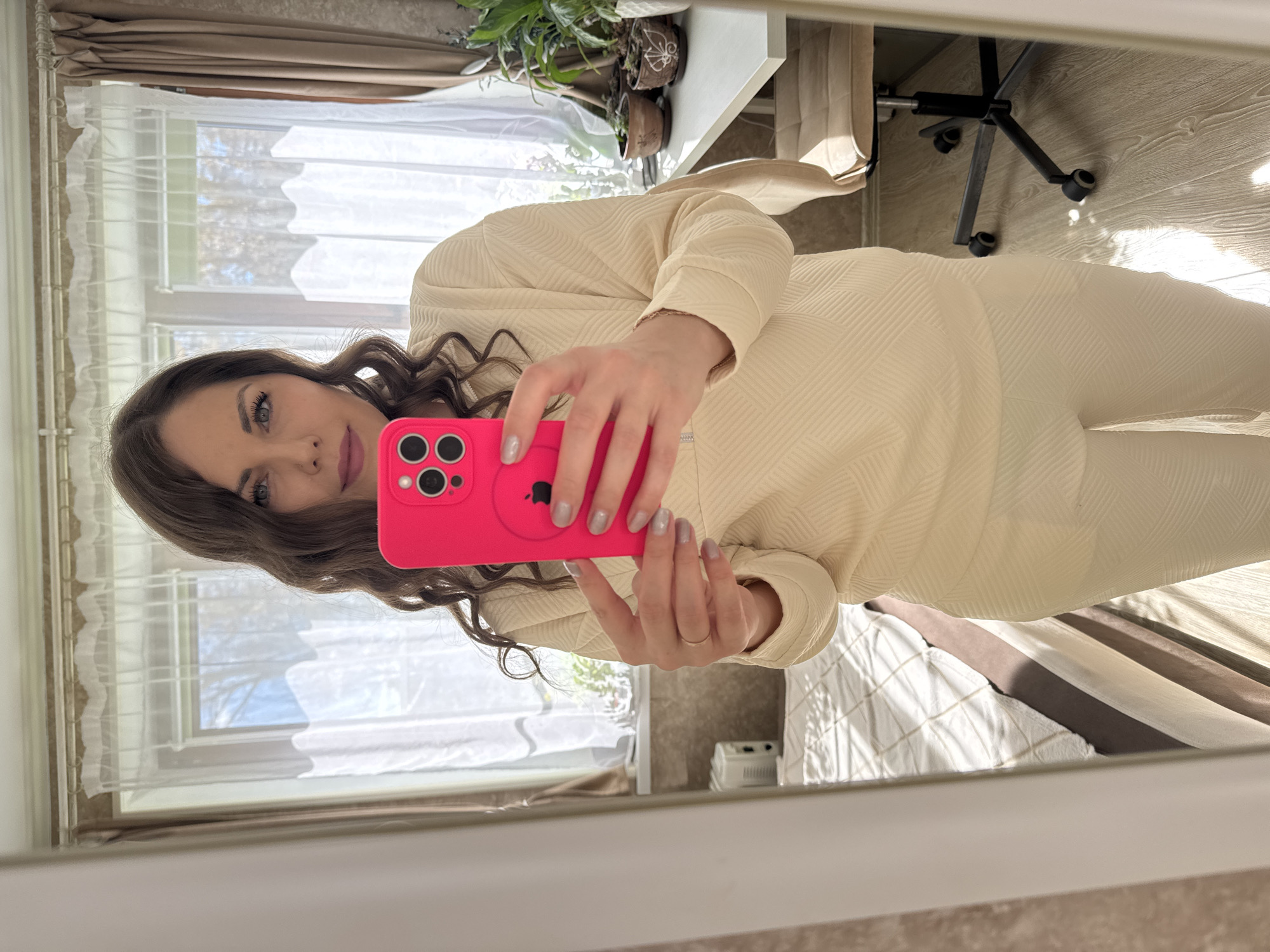 A woman is taking a mirror selfie with a pink smartphone in an indoor setting, with a window visible in the background. A desk and chair are partially visible, along with plants on the desk. The specific location is not discernible from the image.