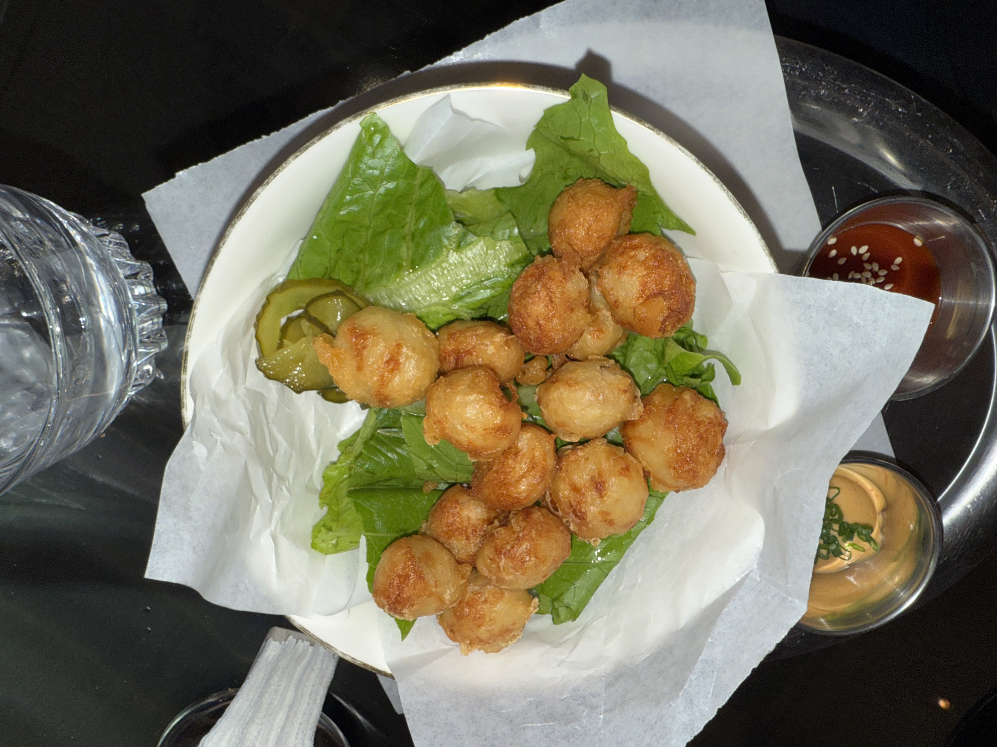 An overhead shot captures a serving of golden-brown fried food presented in a white bowl with a gold rim. The bowl is lined with crumpled white parchment paper and several vibrant green lettuce leaves, upon which the fried items rest. To the left, a small cluster of sliced pickles adds a contrasting green and slightly acidic element.
The fried pieces are small, roughly spherical or irregularly shaped, and appear to be breaded or battered, suggesting they could be anything from fried cheese balls, potato croquettes, or perhaps a type of seafood like scallops or shrimp fritters. Their surface is visibly textured and golden-brown from frying.
Beside the main bowl, on a dark, slightly reflective surface that appears to be a metallic tray, are two small stainless steel ramekins containing dipping sauces. The top ramekin holds a reddish sauce, possibly a sweet chili or sriracha-based sauce, garnished with white sesame seeds. The bottom ramekin contains a creamy, orange-hued sauce, likely mayonnaise-based, topped with finely chopped green scallions or chives.
To the upper left of the main dish, a clear glass with a textured, ribbed base, filled with water, is partially visible. The overall setting appears to be indoors, possibly a restaurant or bar in Yerevan, Armenia, given the dark background and the presentation of the food and drinks. No people are visible, and there is no discernible text in the image.