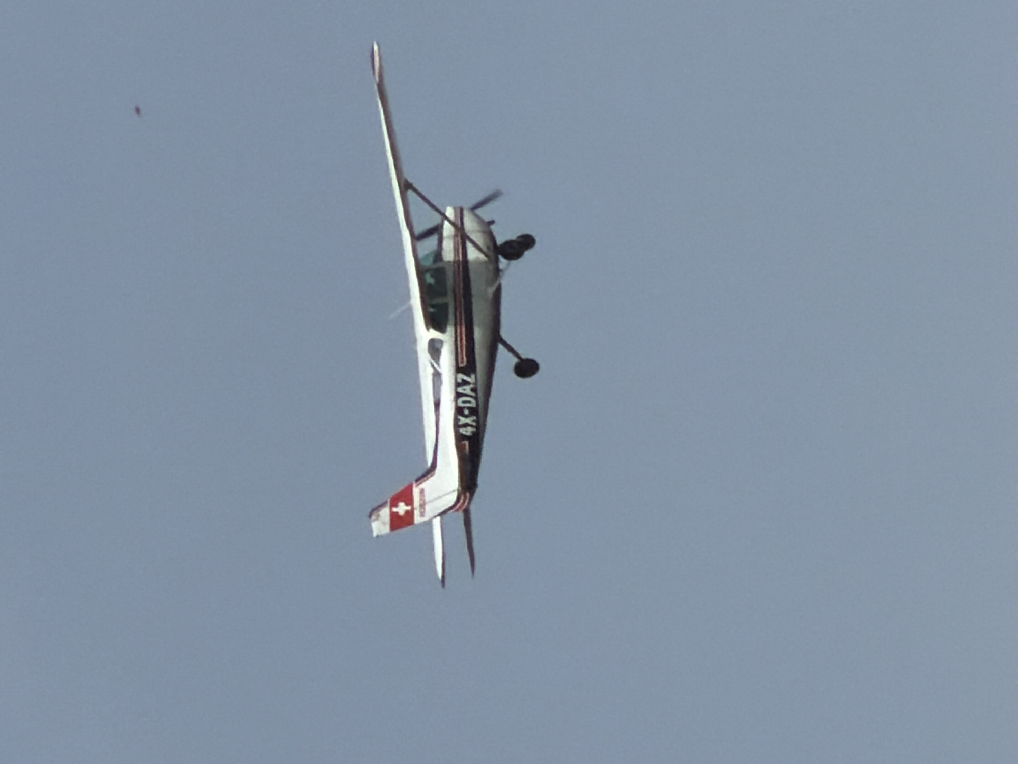 An aerobatic aircraft with tail number 4X-DAZ is performing an inverted flight maneuver against a clear, light blue sky. The plane has a predominantly white color scheme with dark gray accents. A Swiss flag emblem is visible on its tail. A small, dark object is also present in the sky, likely a speck of dust or a distant object. The plane is flying in the city of Herzliya, Israel.