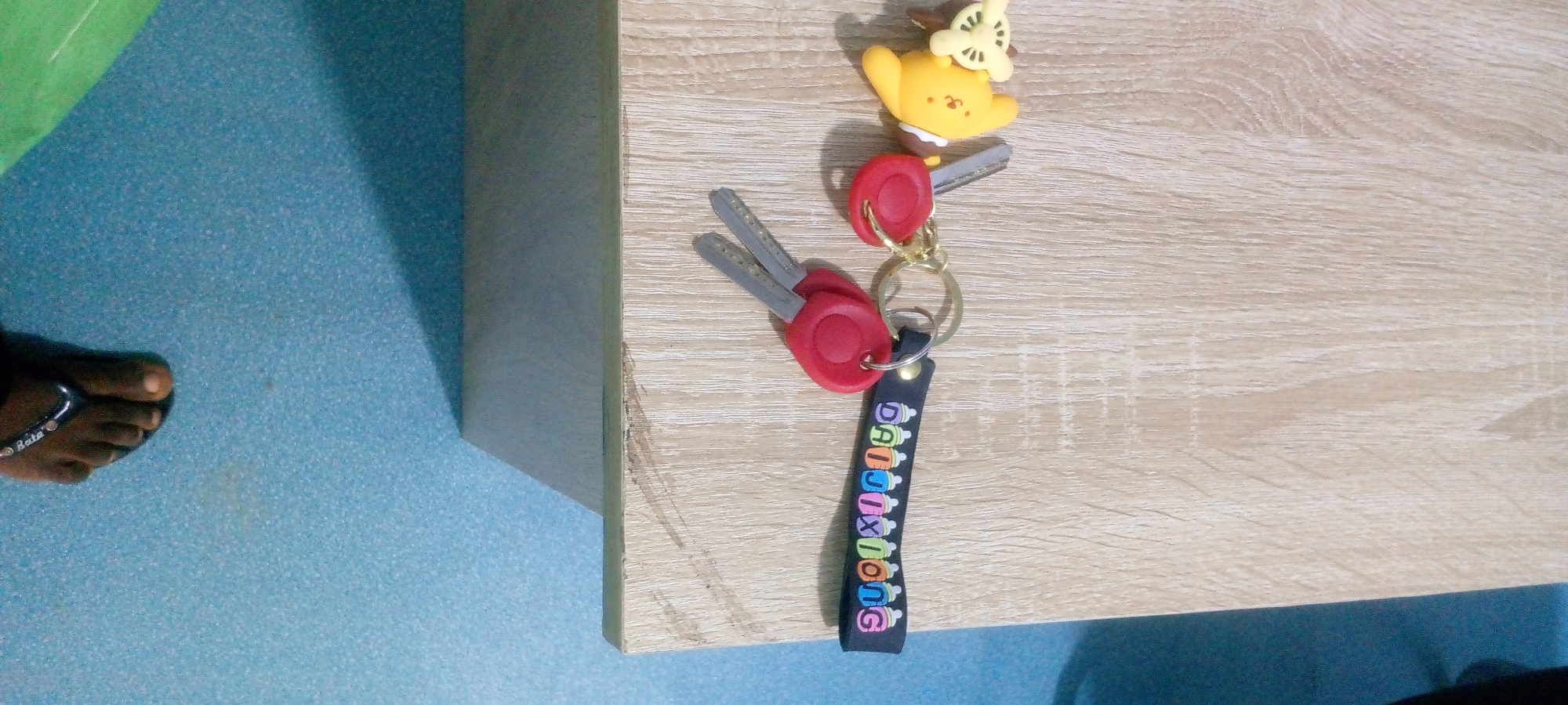 A pair of keys with red key covers and a yellow charm of a character with large ears and small wings are attached to a keychain that has a black fabric strap with the word "DAIJIXIONG" printed on it in colorful letters. The keys and keychain are lying on a light brown wooden surface. To the left of the wooden surface is a blue textured floor with a person's bare foot wearing a black flip-flop on it. The overall scene is an indoor environment, possibly a home or an office, in Indo, Nigeria.