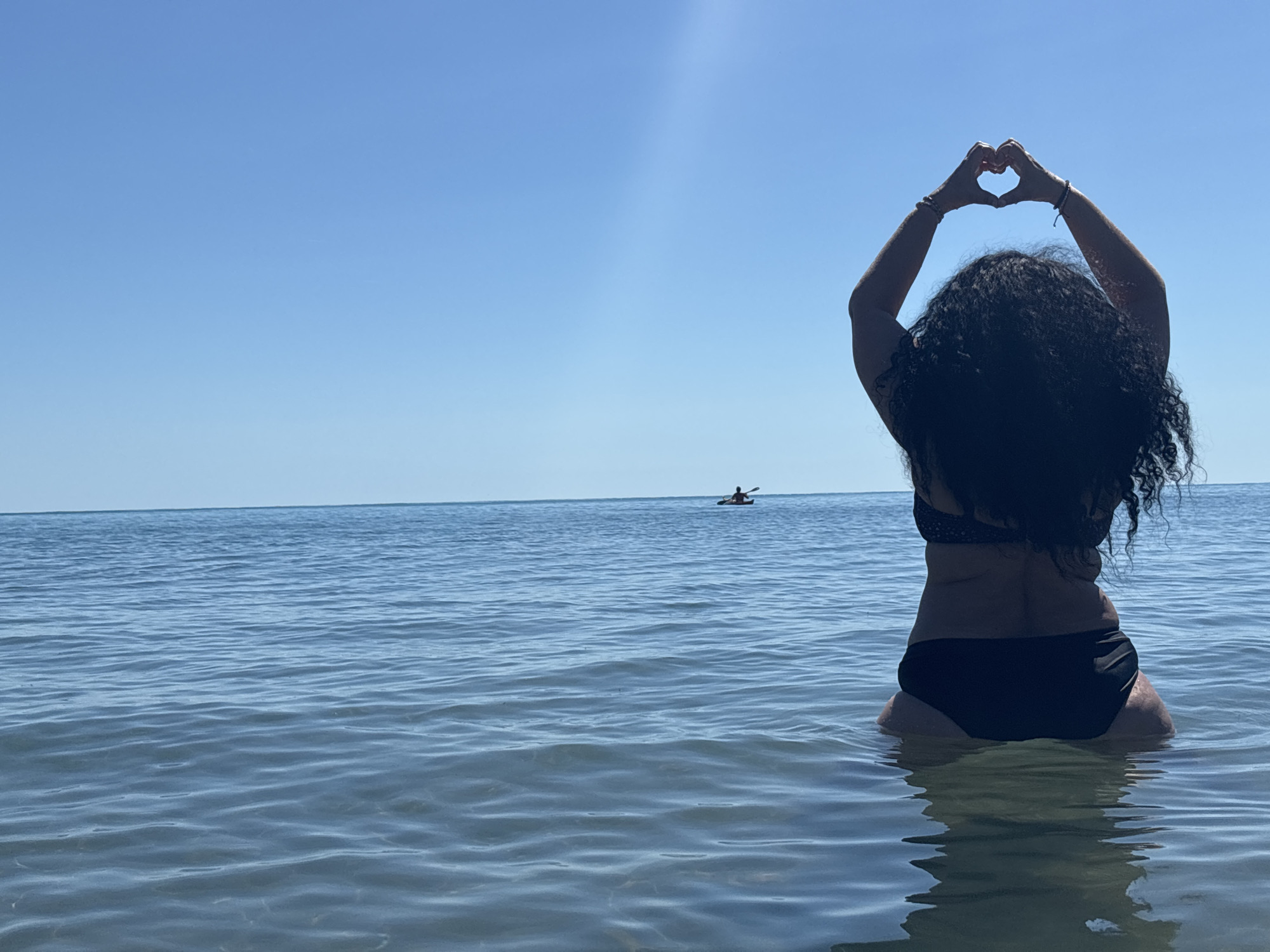 A person wearing a black bikini is sitting in the water at the Black Sea, with their hands forming a heart shape above their head. The person has dark, curly hair. The water is calm and blue, and the sky is clear and bright blue. In the distance, a person is visible in a kayak on the water. The scene appears to be a natural landscape, likely a beach or coastline. The location is identified as Rezovo, Bulgaria.