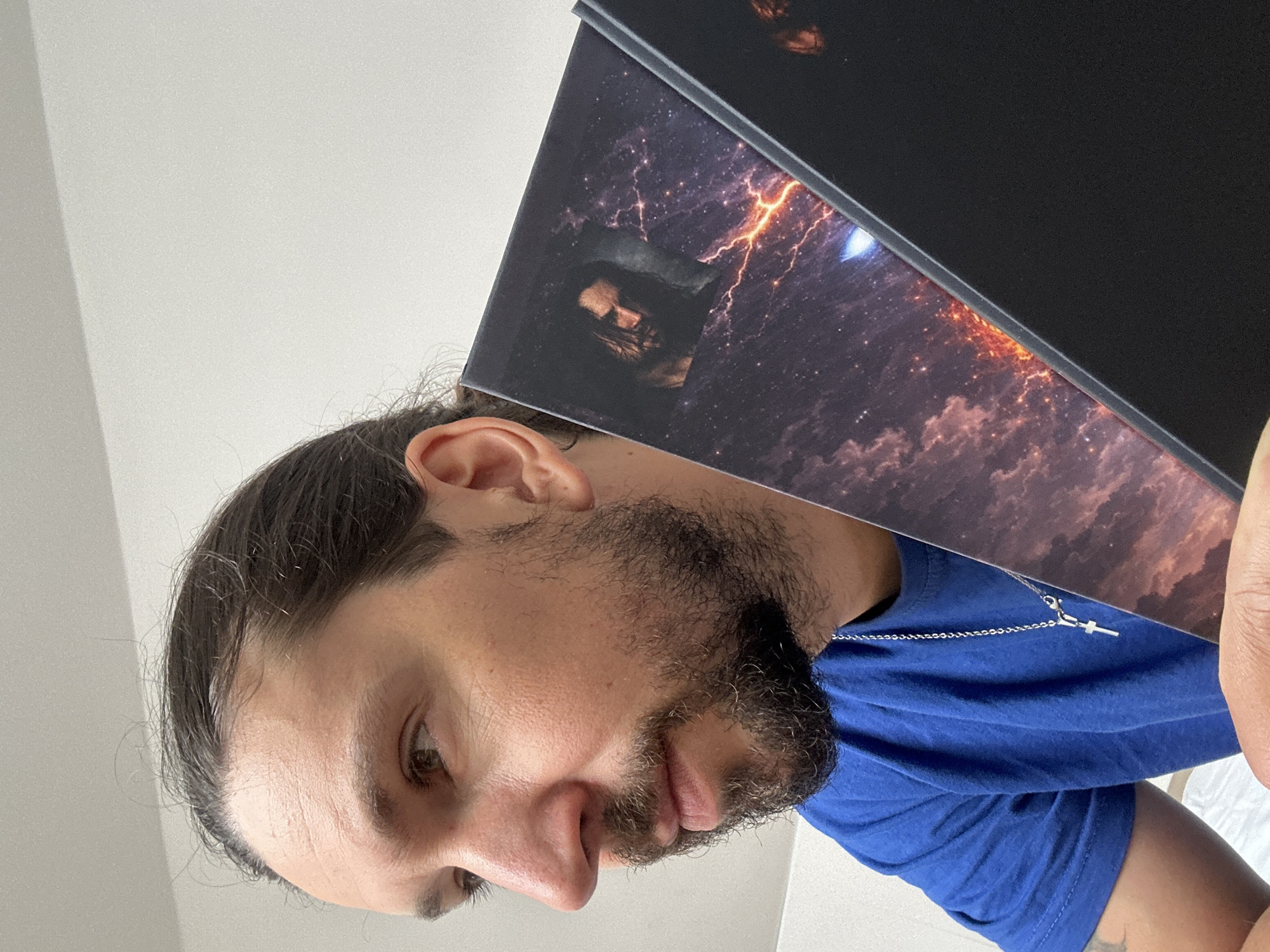 A man with dark hair pulled back into a bun, a beard, and wearing a blue shirt with a silver cross necklace is holding a book with a black cover featuring an image of a figure against a stormy sky with lightning. The background is a plain, light-colored wall. There is no evidence in the image to suggest the location is Nivelles, Belgium.