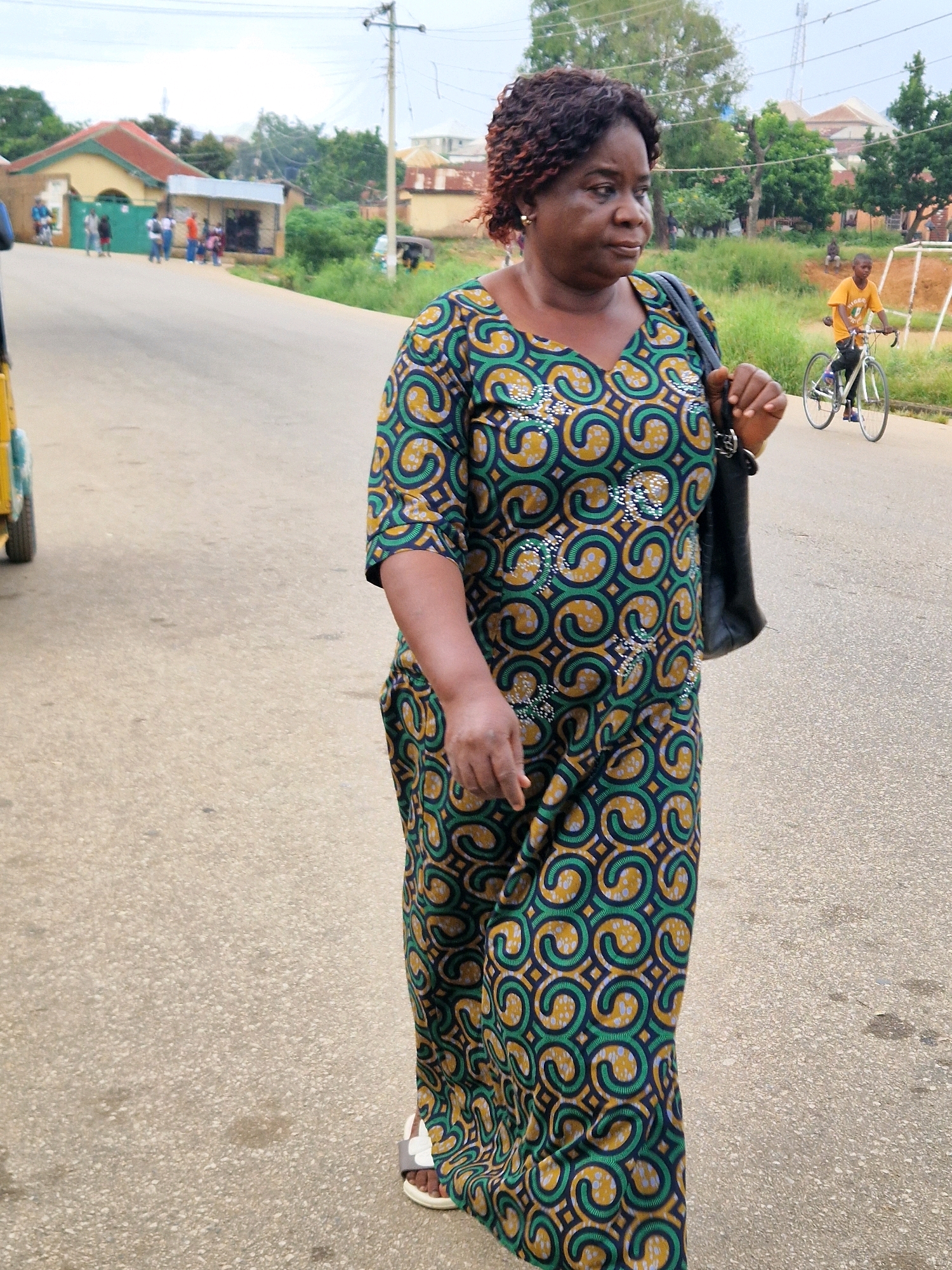 A medium shot captures a woman in a colorful, patterned dress walking along a paved road in Kabong, Nigeria. The woman, who appears to be middle-aged and of African descent, has dark, curly hair and is carrying a black bag over her left shoulder. She is wearing white sandals. To her right, a young boy in an orange shirt is riding a bicycle. In the background, there are buildings, trees, and a utility pole with electrical wires. A yellow vehicle, possibly a tuk-tuk, is partially visible on the left side of the image. The sky is overcast.