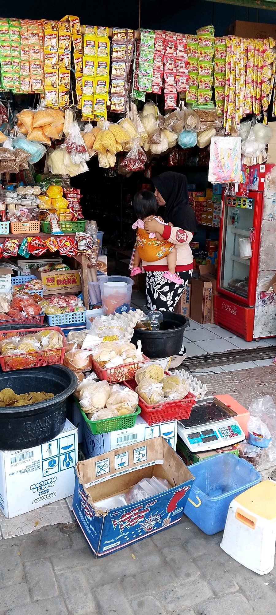 This is a full shot, eye-level, exterior, natural light, medium-distance photograph of a street vendor's stall in Sriamur, Indonesia. The stall is laden with various goods, including bagged snacks, packaged noodles, and other items hanging from a structure overhead. In the foreground, stacked boxes and plastic containers hold more packaged snacks and produce. A woman wearing a black hijab and a patterned skirt is holding a baby wrapped in an orange sling. The baby is facing away from the viewer. To the right of the scene, a red refrigerator stands open, displaying a few items inside. A digital scale is visible on a counter to the right of the stacked boxes. The ground is paved with grey bricks.