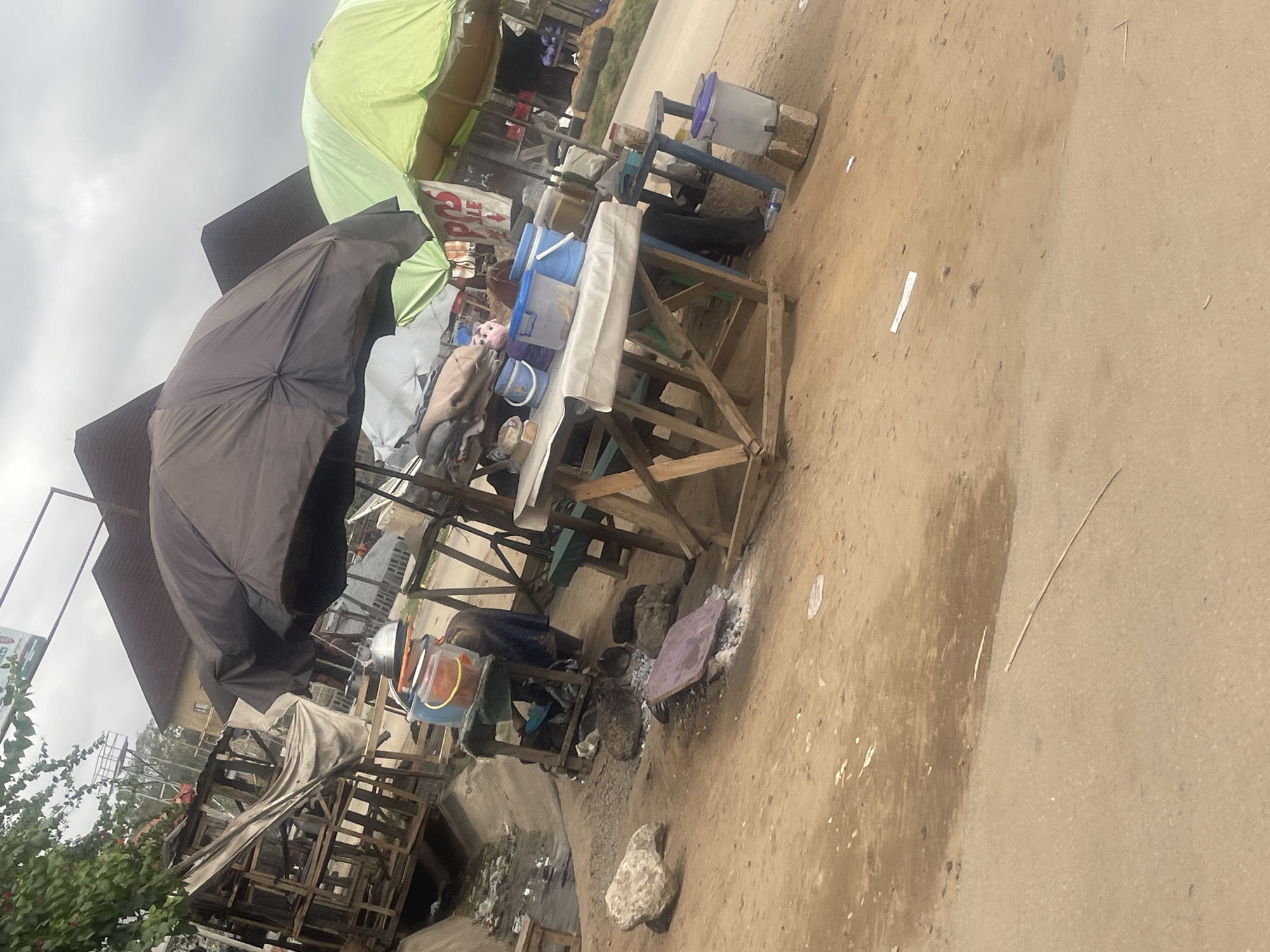 The image depicts a street scene in Abuja, Nigeria. The focus is on a roadside vending stall constructed of wood. The stall is laden with various items, including covered food containers and buckets, and is shaded by a large black umbrella. A small table with a white cloth sits adjacent to the stall.
To the left of the stall is a dilapidated wooden structure filled with trash. The street is made of dirt and is littered with small pieces of debris. The sky is overcast. No people are clearly visible.