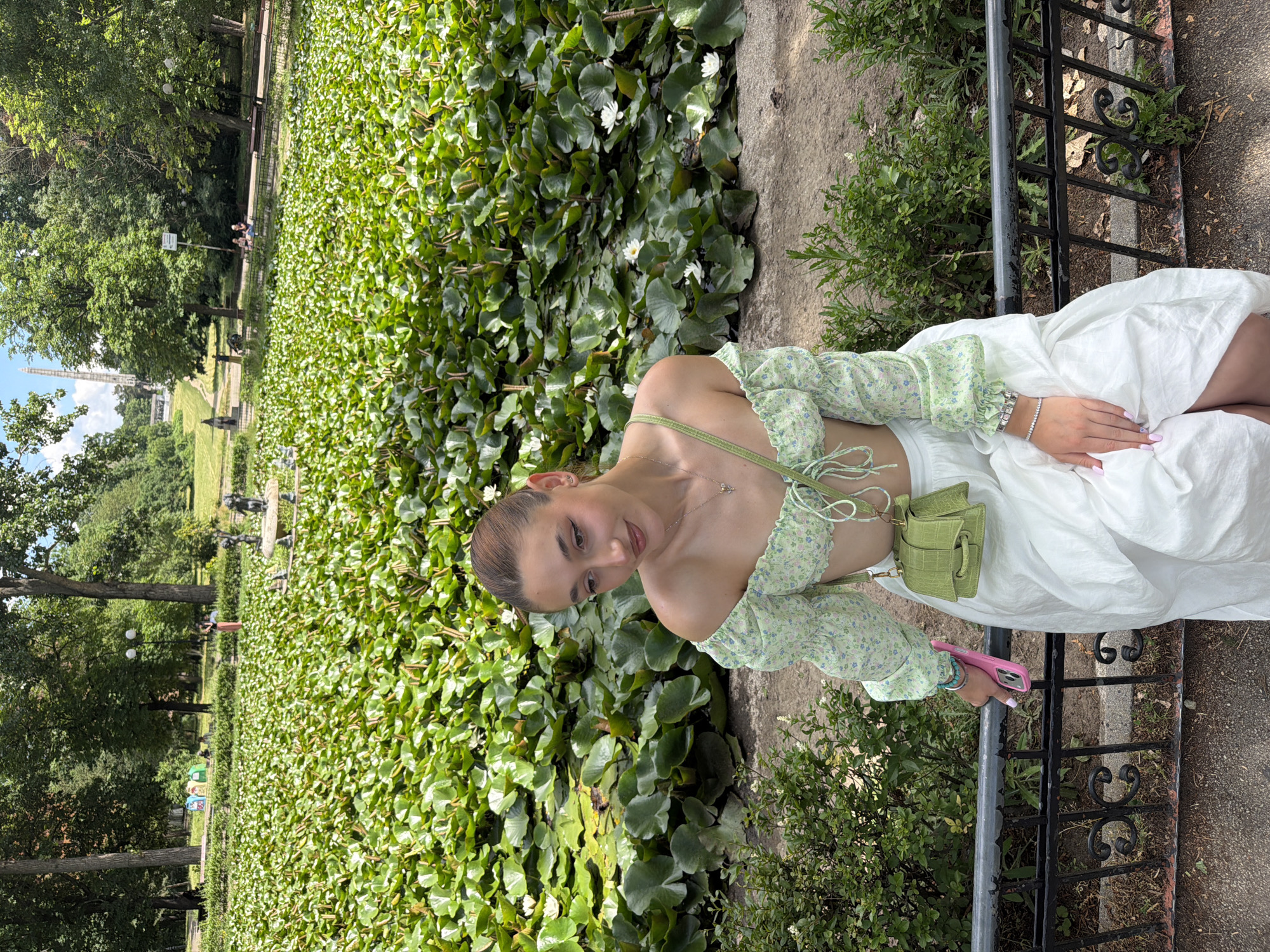 A young woman with brown hair pulled back, wearing a green floral off-the-shoulder crop top and a white maxi skirt, is seated outdoors. She is wearing a small green crocodile-textured crossbody bag and a silver bracelet on her left wrist. In her right hand, she holds a pink phone. She is leaning on a black metal fence, with a dense area of water lilies and lily pads behind her. The background shows a park with trees, walking paths, and other people in the distance. The scene appears to be in the Borisova Gradina park in Sofia, Bulgaria, specifically near the lake with water lilies. The lighting suggests it is daytime.