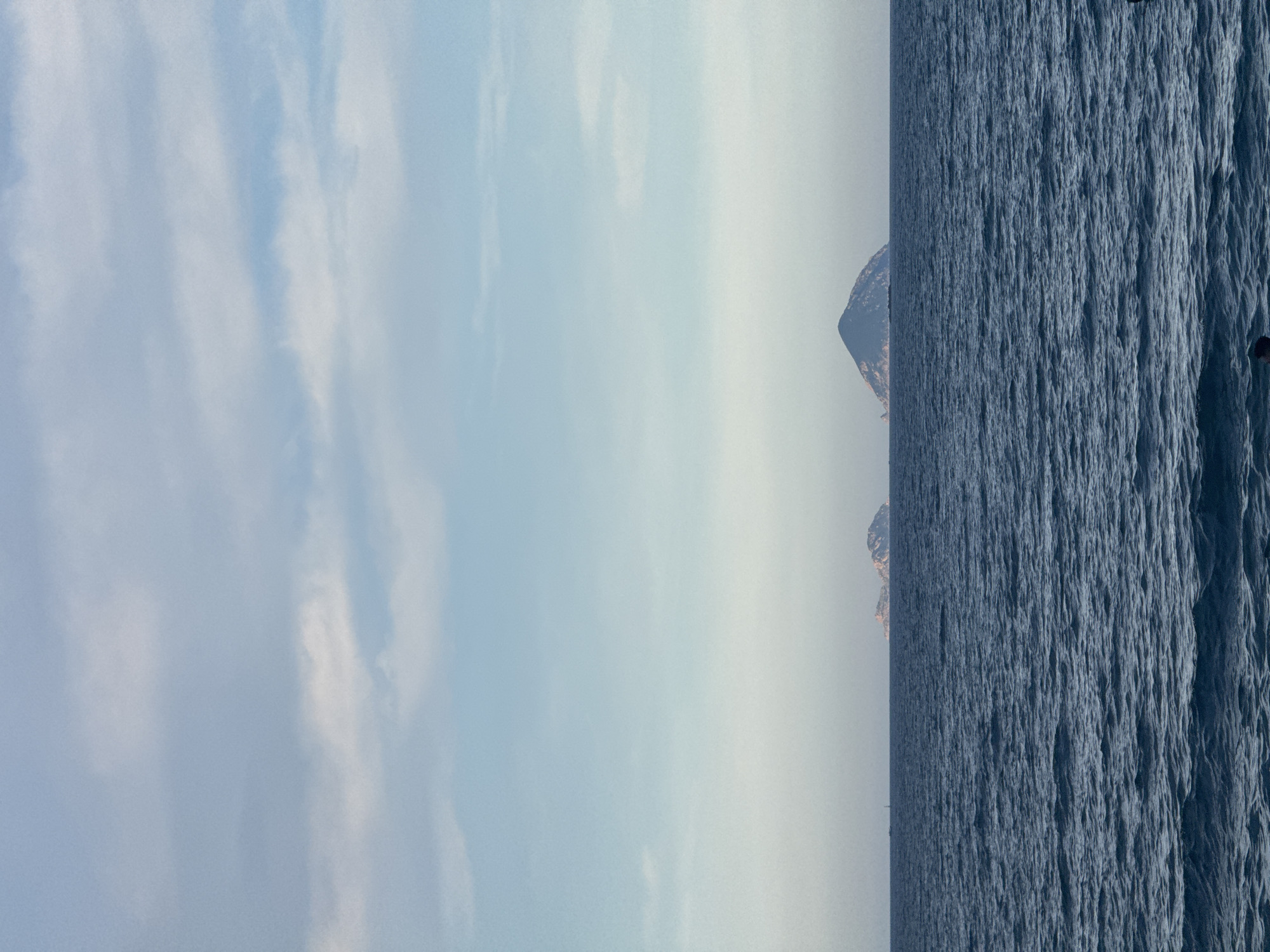 This image captures a serene seascape, possibly near Nha Trang Ward, Vietnam. The scene is dominated by the vast expanse of the ocean on the right, its surface rendered in a deep, textured blue, suggesting a slightly choppy sea. The water stretches to the horizon, where it meets a hazy sky.
On the left side of the frame, a soft, pale blue sky transitions into lighter tones towards the horizon, with delicate white clouds scattered across it. The clouds appear wispy and form elongated streaks, indicative of a clear or partly cloudy day. In the distance, silhouetted against the hazy sky, are the faint outlines of islands or rocky outcrops. The closest visible landmass appears as a dark, rugged shape, partially illuminated by the sun.
The time of day is suggested to be late afternoon or early morning due to the soft light and the hazy atmosphere, which often occurs during these periods. The overall mood is tranquil and expansive, evoking a sense of peace and the raw beauty of nature. There are no discernible people, objects, or text within the visible frame. The image presents a natural landscape, likely a coastal view, emphasizing the interplay between sky, sea, and distant land.