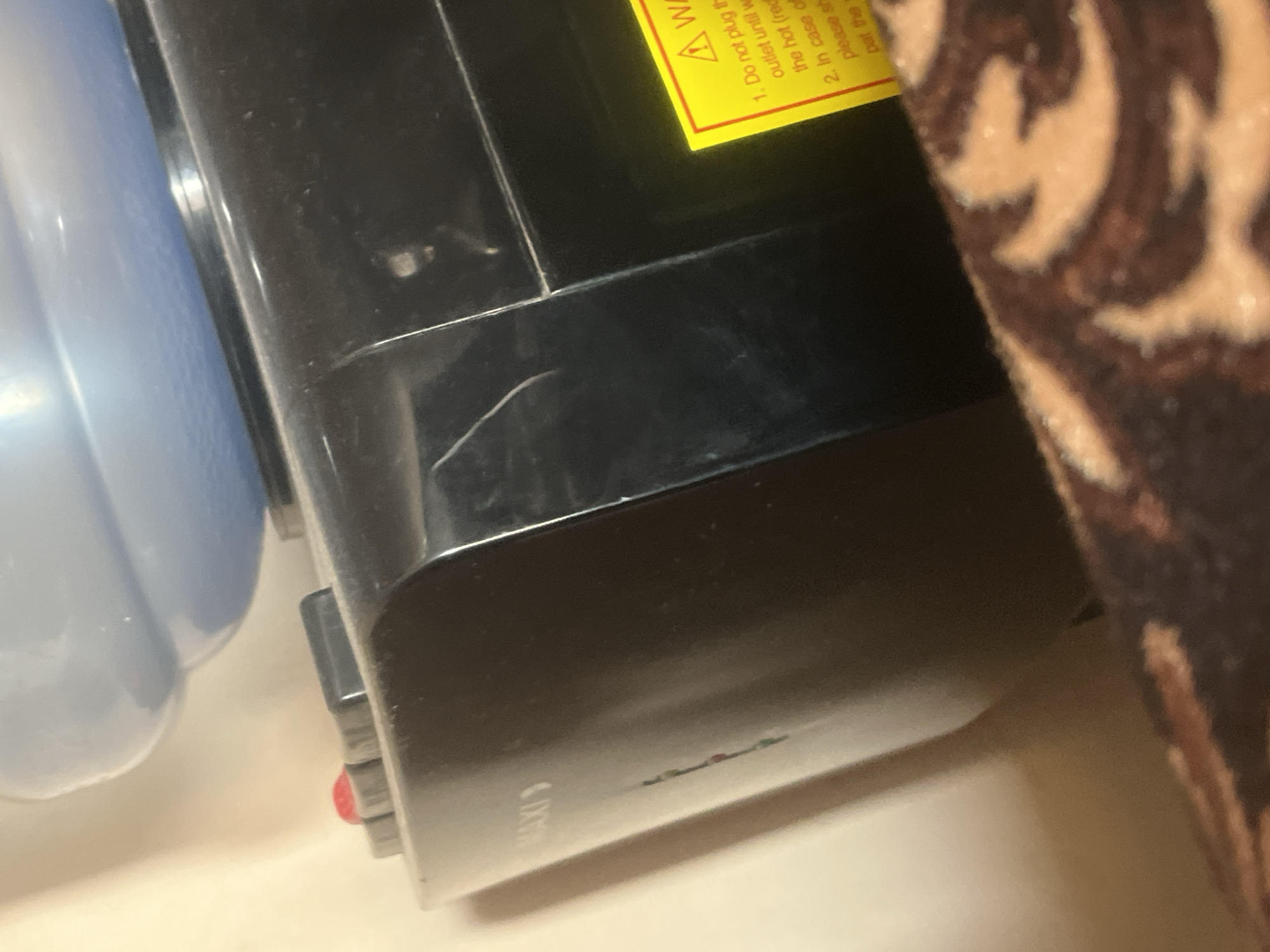 The image shows a close-up of a black water cooler dispenser. A blue water bottle is visible at the top. On the front of the dispenser, there are two buttons, one red and one gray. A yellow warning sticker with text in English is affixed to the right side of the dispenser. The text on the sticker appears to be instructions for use and safety precautions. The dispenser is placed against a cream-colored wall. The foreground of the image is occupied by a textured brown and beige fabric, possibly a blanket or upholstery. The image does not provide any information about the location being in Jauro Yinu, Nigeria, or any details about people or actions.
