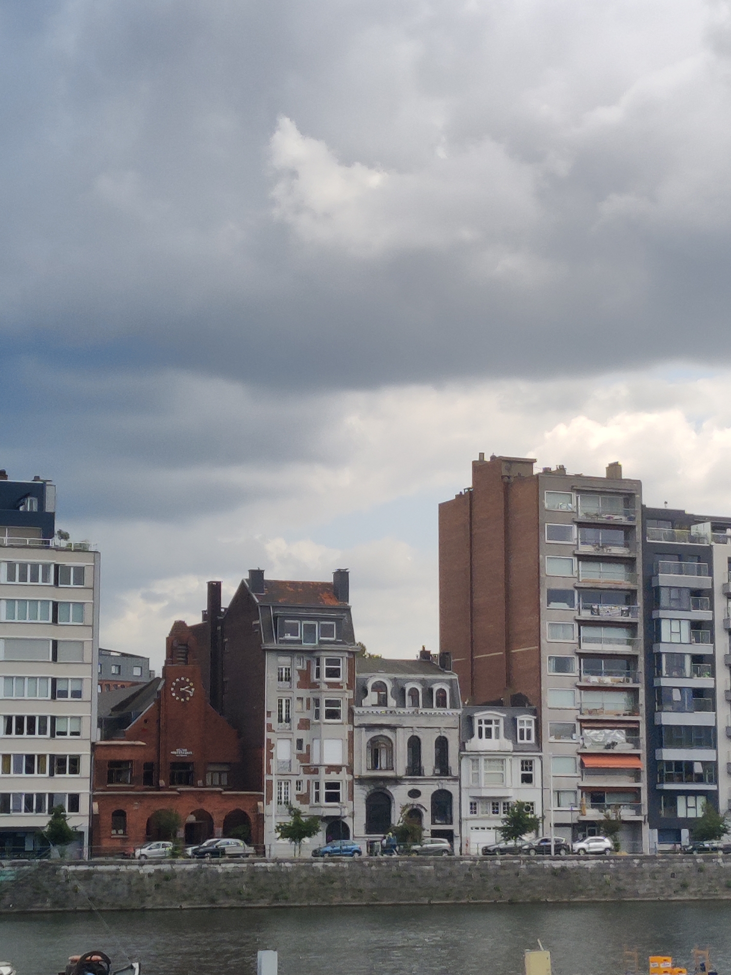 The image shows a waterfront view of buildings in the city of Liège, Belgium. The architecture varies from modern high-rise apartments to older, more ornate structures. The buildings are situated along a river or canal, with a gray concrete embankment in the foreground. Several cars are parked along the street in front of the buildings. The sky is overcast with dark clouds, suggesting a potential for rain. The overall impression is of an urban waterfront with a mix of architectural styles.