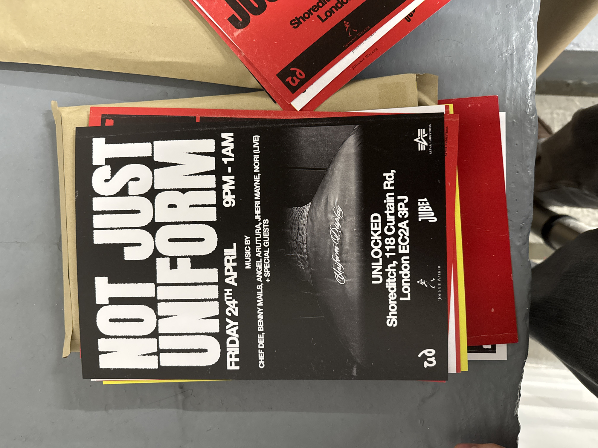 An overhead shot captures a stack of promotional flyers and cards resting on a dark grey, textured surface. The most prominent item is a black flyer for an event titled "NOT JUST UNIFORM," displayed in large white block letters.
The black flyer details a music event scheduled for "FRIDAY 24TH APRIL" from "9PM - 1AM." It lists musical acts including "CHEF DEE, BENNY MAILS, ANGEL ARUTURA, JHERI MAYNE, NORI (LIVE) + SPECIAL GUESTS." The event location is specified as "UNLOCKED Shoreditch, 118 Curtain Rd, London EC2A 3PJ," placing it within London, United Kingdom. An obscured black and white image on the flyer shows a person's torso, possibly wearing a jacket, with the text "Uniform Display" overlaid.
Beneath the main flyer, a mix of red, white, and yellow cards are partially visible. Another set of red cards is stacked at the top of the image, revealing text such as "Shoreditch Londo" (likely London) and "JOHNNIE WALKER." Visible brand logos on the flyers include "JOHNNIE WALKER," "JUBEL," "ALPHA INDUSTRIES," and a stylized "ud" logo. The background features a light brown paper or cardboard surface, with a small portion of dark clothing visible on the far right. The flyers advertise an upcoming night-time event, likely a music party or gig, with multiple sponsors.