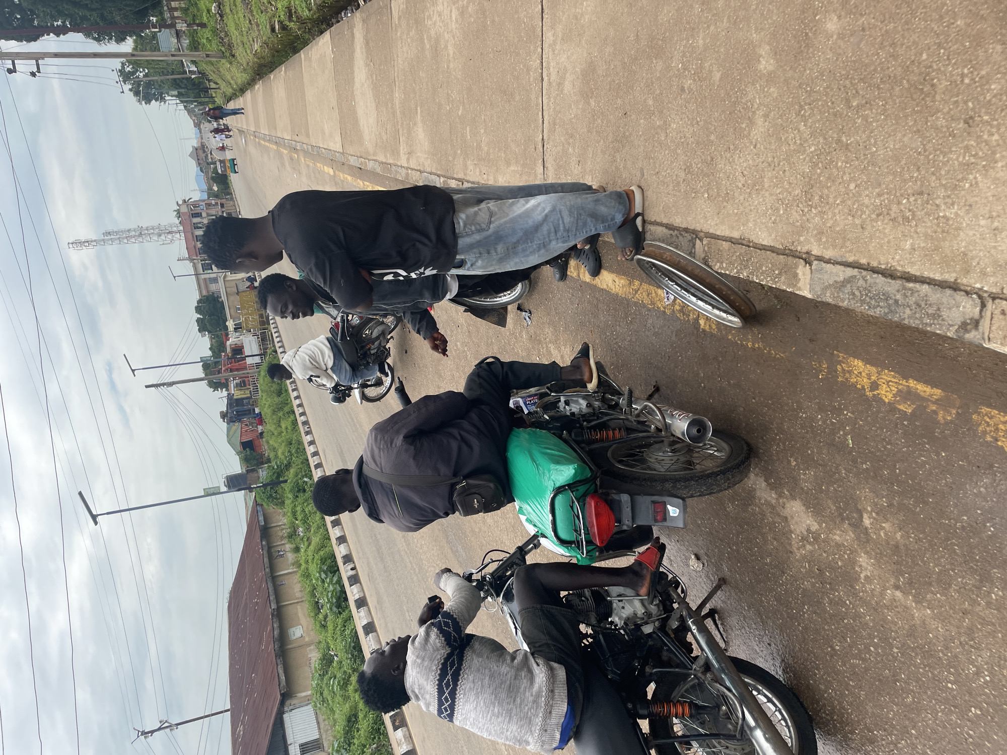 In Jos, Nigeria, three men are gathered on the side of a road next to motorcycles. Two men are seated on motorcycles, while a third man stands. One of the seated men wears a grey sweater and shorts, with red sandals. The other seated man is dressed in a dark jacket and appears to be talking with the standing man, who is wearing a black t-shirt and light blue jeans. Behind them, another man sits on a motorcycle. The motorcycles are parked on a paved road with a yellow line marking the lane. A concrete sidewalk runs alongside the road, and a grassy area with vegetation is visible beyond the sidewalk. In the background, buildings, power lines, and communication towers are present. A detached motorcycle wheel lies on the ground near the standing men. The sky is overcast, suggesting a cloudy day.