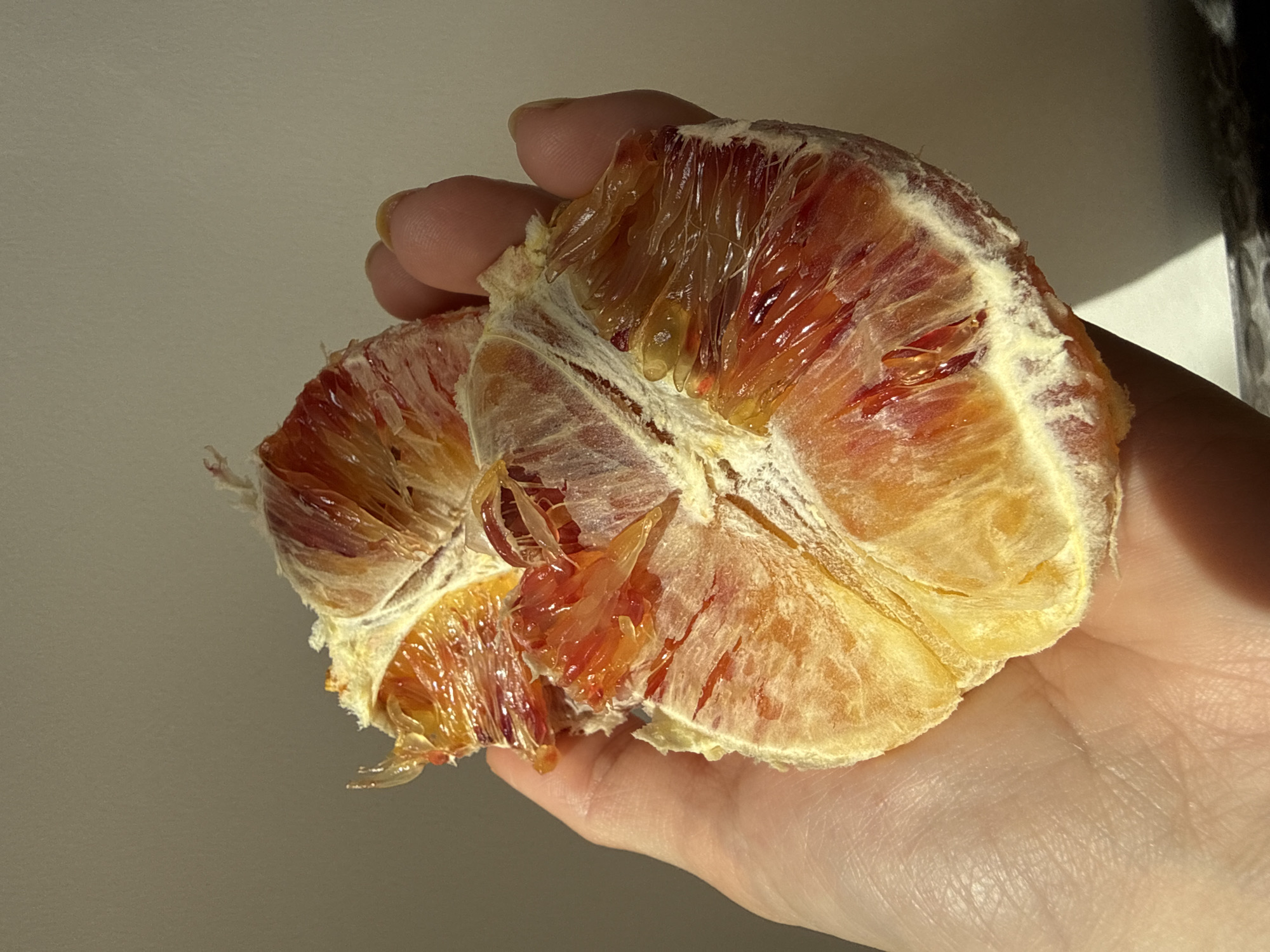 A close-up image shows a hand holding a blood orange that has been peeled. The blood orange segments are visible. The hand appears to be peeling the orange. No location information is discernible from the image.