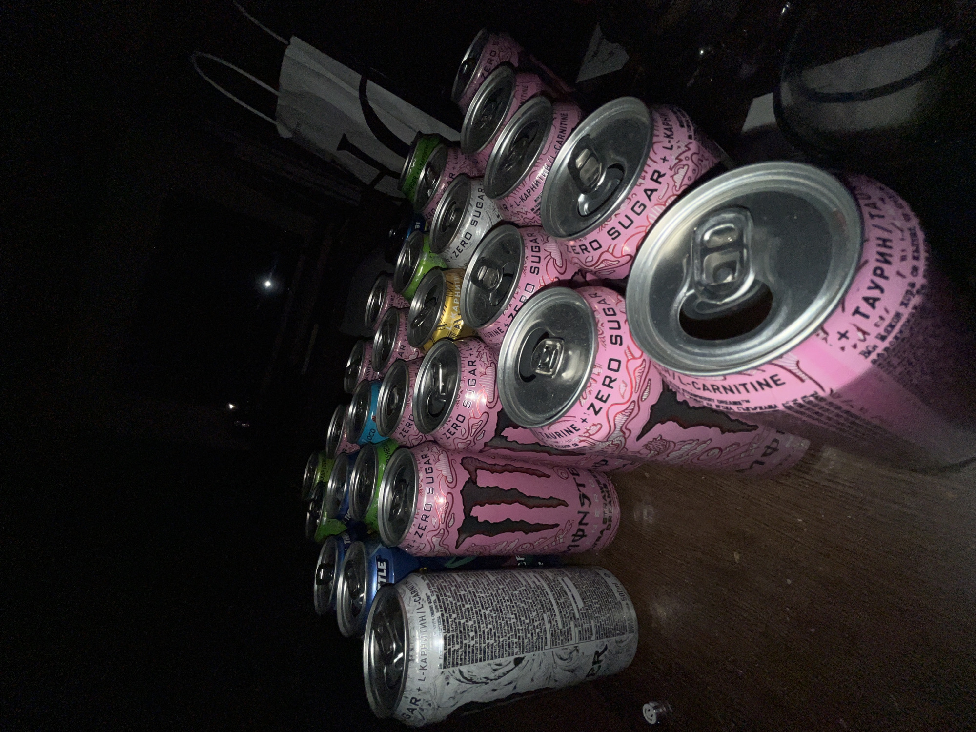 The image displays approximately 25-30 beverage cans, primarily energy drinks, resting on a dark, horizontal surface, possibly wood, within a dimly lit indoor environment in Sofia, Bulgaria.
The majority of the cans are pink Monster Energy "Zero Sugar Ultra Strawberry Dreams" flavor, identifiable by their color, distinct claw logo, "MONSTER" branding, and "ZERO SUGAR" text. Many of these pink cans have open pull tabs, indicating consumption. A white 500ml Monster Energy can, featuring extensive Cyrillic text including "L-КАРНИТИН / L-CARNITINE," is prominently positioned on the left side. Several other energy drink cans in blue, green, yellow, and silver are intermingled among the pink and white cans; some of these also display Cyrillic labeling, such as "ЗАХАР / ZER" on a silver can. The cans are arranged in a dense, somewhat disorganized cluster across the surface.
The supporting surface is dark brown or black, with minor dust particles visible. The background is predominantly dark, almost black, with faint light reflections and an indistinct white object resembling a shopping bag or part of a furniture item visible in the upper right quadrant. No individuals are present in the scene.