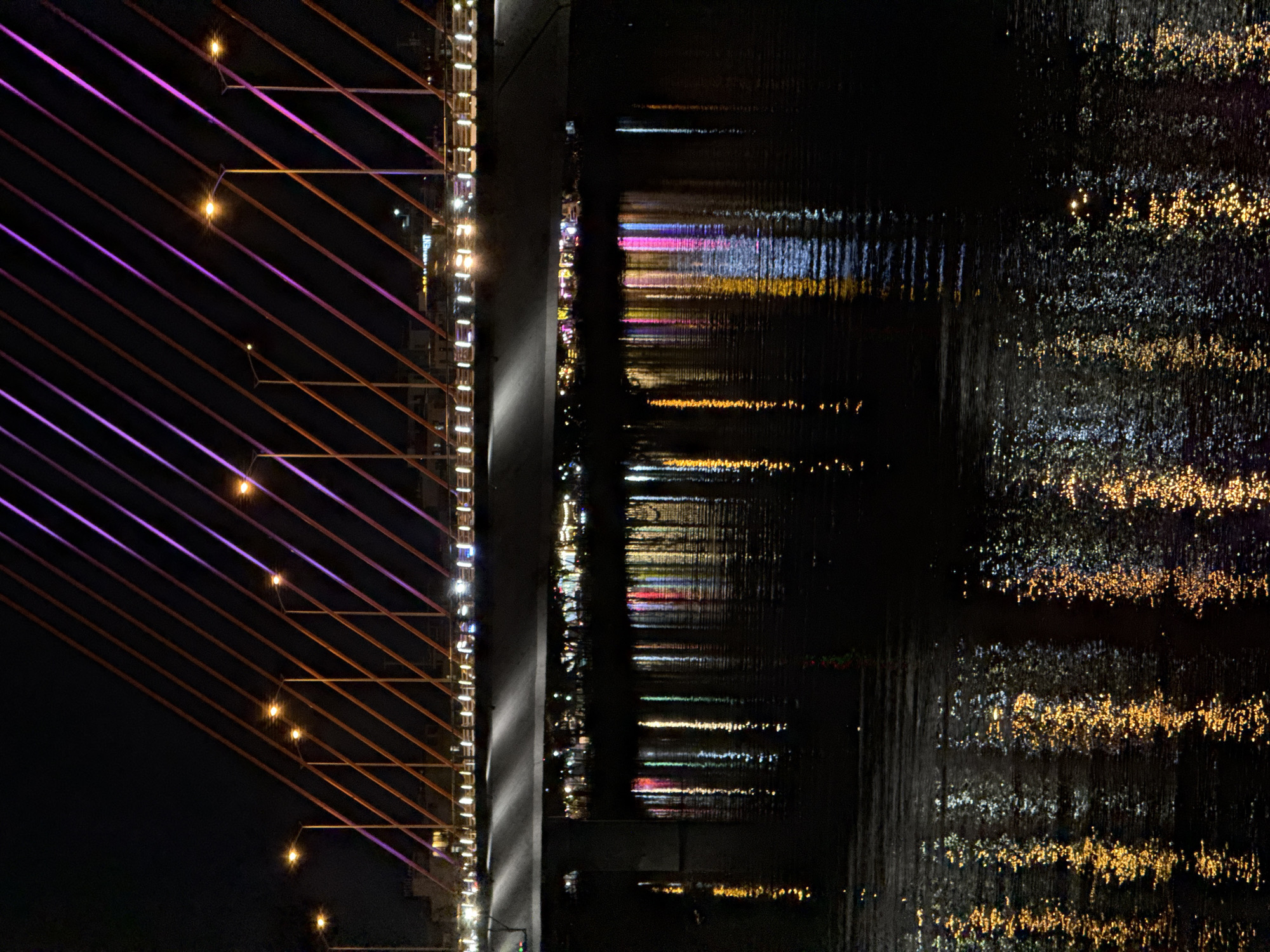 The image captures a nighttime scene of the Han River in Da Nang, Vietnam, featuring the impressive Dragon Bridge. The bridge's suspension cables are illuminated with alternating bands of orange and purple light, creating a striking visual against the dark night sky. Several streetlights are visible along the bridge's roadway, casting warm yellow glows. Below the bridge, the river's surface reflects the vibrant lights from the bridge and the cityscape, creating a shimmering tapestry of colors, including streaks of yellow, pink, and blue. The water is dark, with the reflections appearing as elongated, distorted patterns. The overall atmosphere is one of urban nighttime beauty and tranquility.