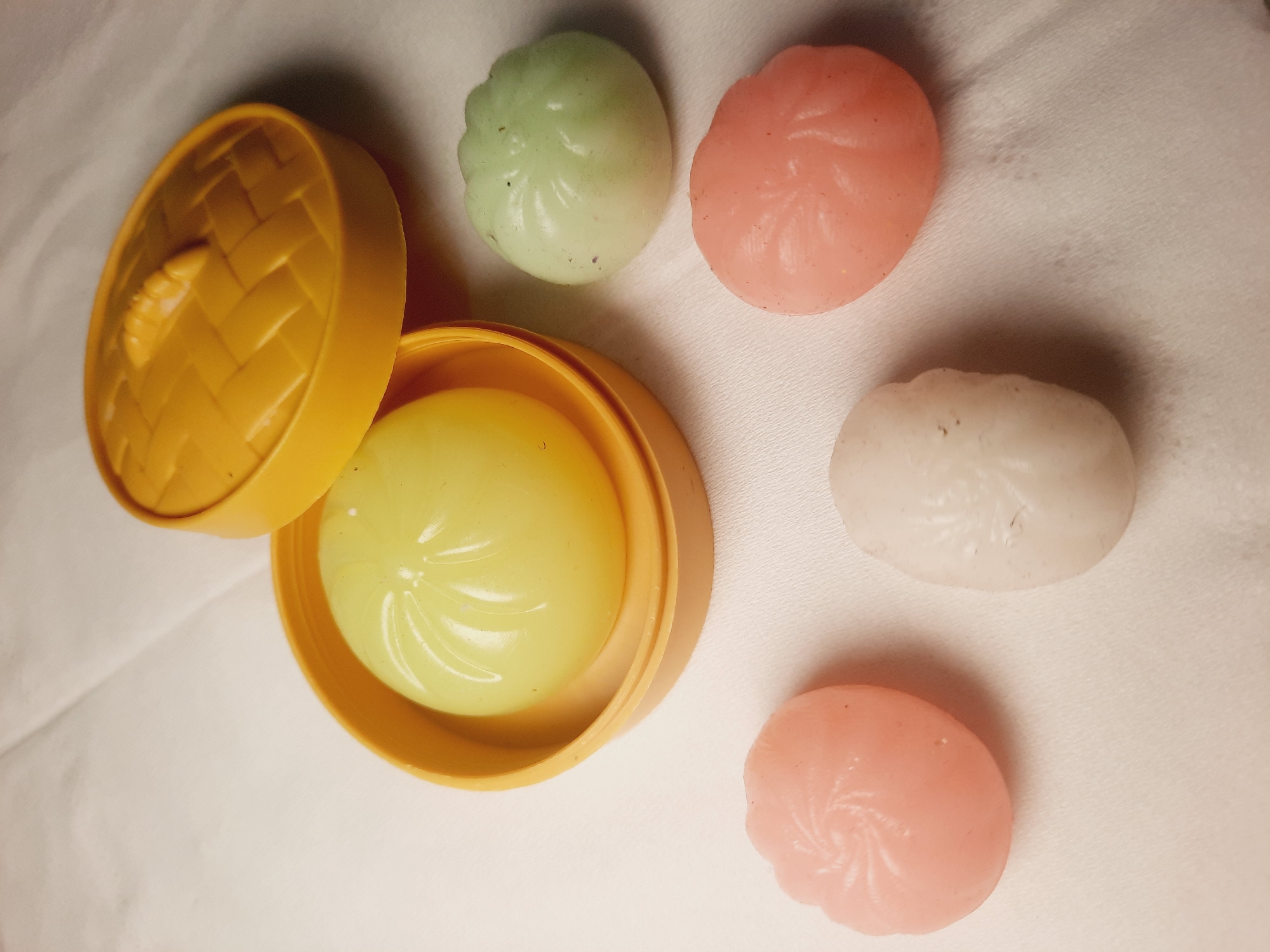 A yellow plastic container, resembling a steamer basket, holds a light yellow, bun-shaped object. The lid of the container, also yellow and textured to look like woven material, sits beside it. Scattered around the container are four other bun-shaped objects: one light green, two pink, and one white. The objects are resting on a white, textured surface, possibly fabric. There are no people visible in the image.