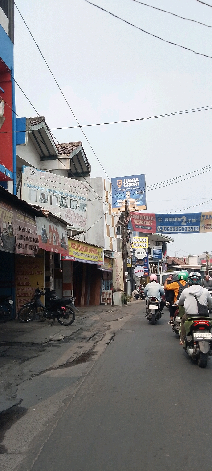 This image depicts a street scene in Karangsatria, Indonesia, likely during daylight hours under an overcast sky. The foreground shows a cracked and weathered asphalt road with some standing water and debris. On the left side of the road, a sidewalk area features parked motorcycles, including a black motorcycle with a red taillight. Adjacent to the sidewalk are storefronts with various signs, one of which advertises "PUSAT PLAFON PVC & INTERIOR" and mentions "SAHABAT INTERIOR RUMAH ANDA". Another sign appears to list building materials such as "PLAFON PVC", "SEKO 2000", "WALL PANEL", and "LIST PVC".
The middle ground of the image shows several people riding motorcycles traveling down the street. One rider in front wears a light blue shirt and a pink helmet. Behind them, another rider wears an orange shirt and a green helmet, followed by a rider in a light gray hooded jacket and a green helmet. Further down the road, more motorcycles and individuals are visible.
The background is dominated by utility poles and overhead wires, along with various advertisements. A large billboard with the text "JUARA GADAI" and "Cara Juara Kalahkan Masalah" is prominent. Another banner reads "CICILAN 2 JT-AN" with a phone number "0823 2500 3919". Smaller signs and shop awnings are also visible, indicating a commercial area. The buildings appear to be a mix of single and multi-story structures. The overall impression is a busy, ordinary street in an Indonesian town.