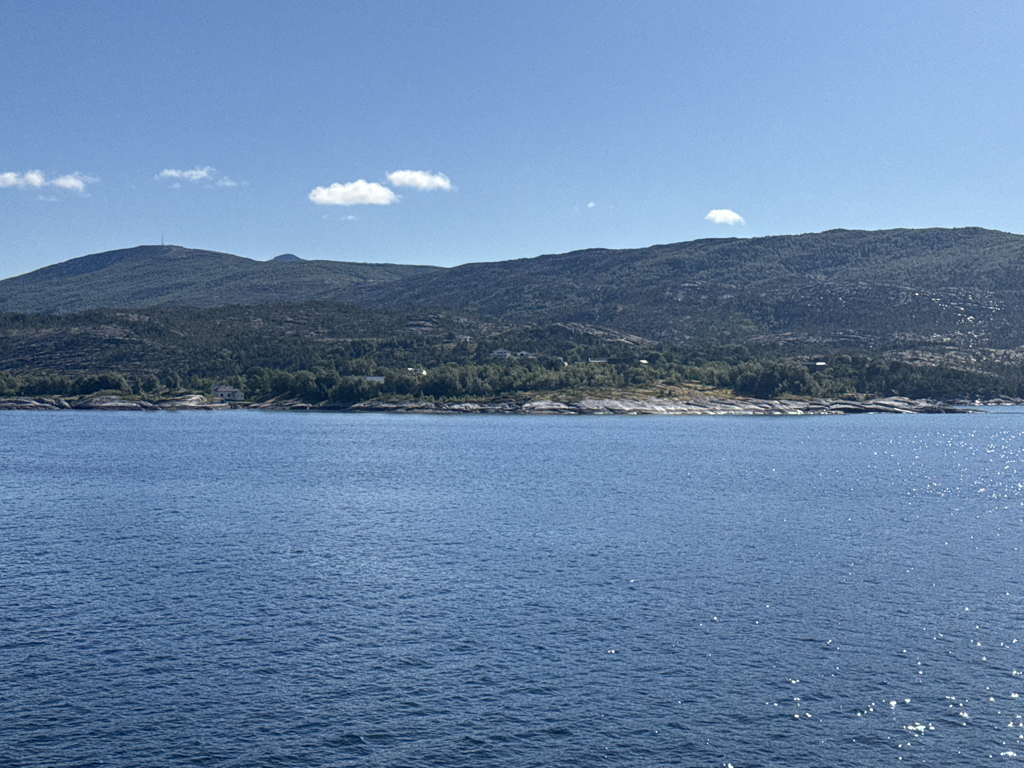 This media file depicts a scenic view of a body of water, likely a fjord or bay, in Bognes, Norway. The foreground is dominated by the expansive blue water, reflecting the sunlight with numerous small, sparkling glints. The water appears calm, with gentle ripples across its surface.
In the background, a verdant, tree-covered hillside rises steeply, forming a natural backdrop. Scattered amongst the trees and along the rocky shoreline are a few scattered houses, suggesting a sparse residential area. The hillside extends to meet a range of rolling mountains in the distance. The highest peak in the left portion of the image features a prominent tower, possibly a telecommunications mast. The sky is a clear, bright blue, with a few small, wispy white clouds scattered across it.
The scene is bathed in bright sunlight, indicating it is daytime, likely midday or early afternoon, given the angle of the light and the clarity of the sky. The weather appears to be pleasant and sunny, typical of a summer day in this region. The overall impression is one of natural beauty and tranquility. There is no visible text within the image.