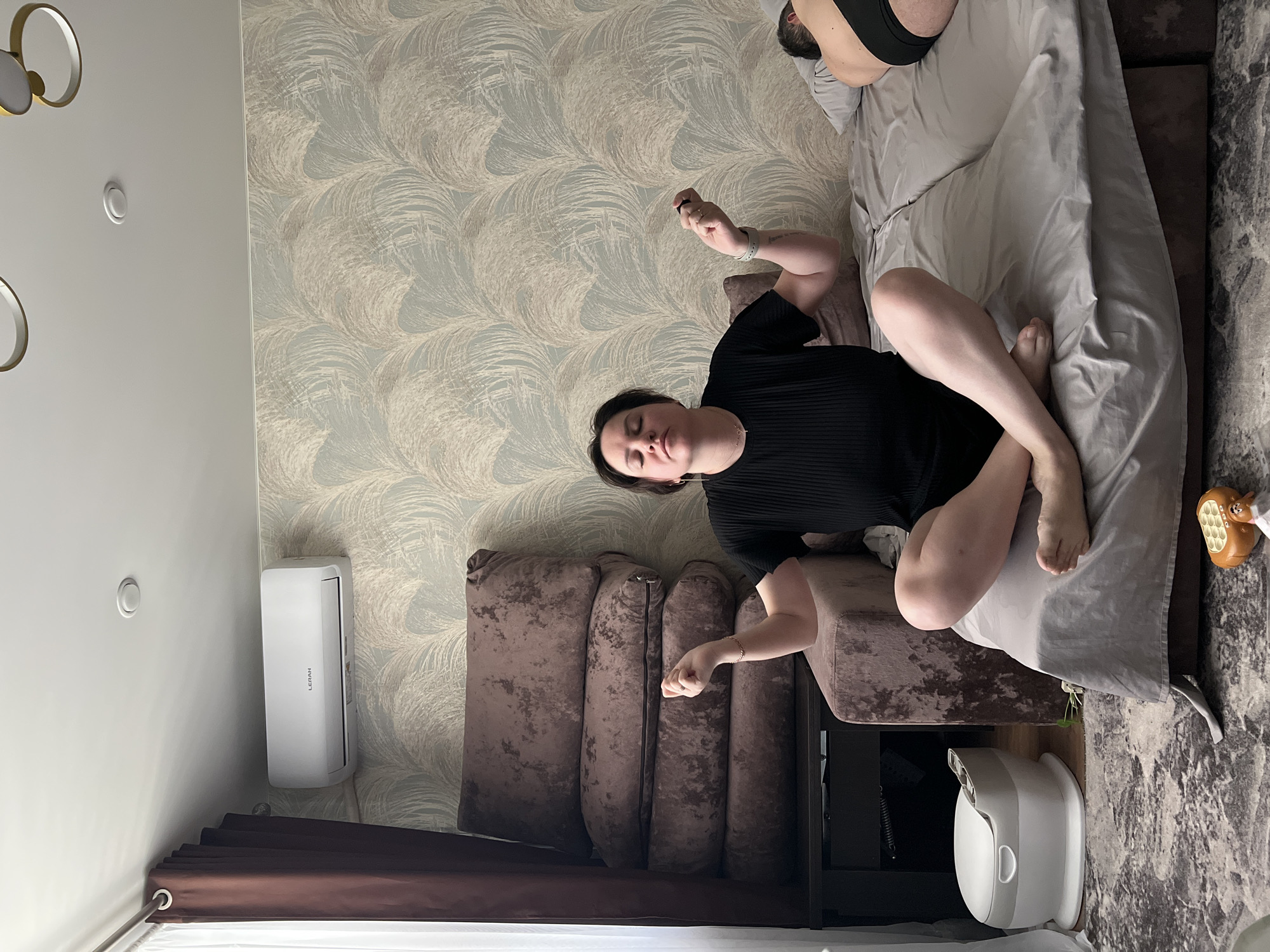 A woman in a black shirt is sitting cross-legged on a brown couch in a room with patterned wallpaper. Another person is lying behind her on the couch. An air conditioner is mounted on the wall to her left. There's a white appliance near the couch. The location is Noviy, Russia.