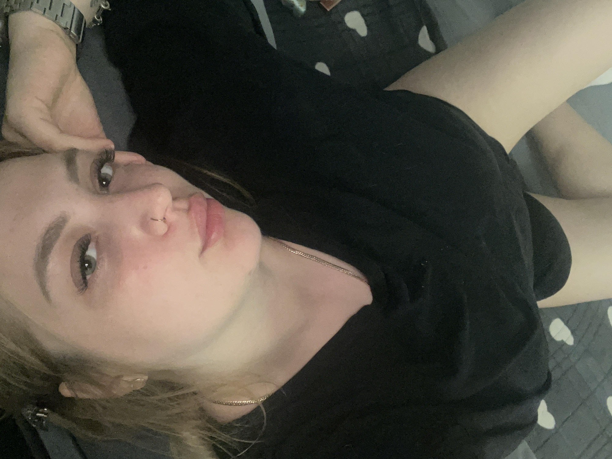 A woman is lying down. She wears a black shirt and a metal watch. She has a nose ring and a gold chain around her neck. She rests a hand near her head. The background is a gray blanket with white heart shapes. There is no indication of the location being Izhevsk, Russia.