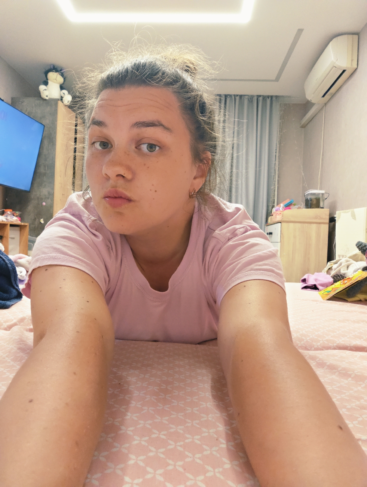 The image depicts a female individual, appearing to be an adult, lying prone on a bed and looking directly into the camera. The scene is set in a residential bedroom in the city of Chaikovsky, Russia. The individual has light skin with visible freckles on her face and forearms. Her light-colored hair is gathered into a high bun or topknot, with multiple loose strands around her head. She is wearing a light pink short-sleeved t-shirt, and a small gold-colored hoop earring is visible in her left ear. Both forearms are extended towards the camera, indicating she is holding the photographic device. Her lips are slightly pursed.
The bed is covered with a light pink fitted sheet featuring a repeating white geometric pattern resembling clover shapes. The room's walls are light-colored, and the white ceiling is equipped with a linear recessed light fixture and a rectangular decorative recess. An air conditioning unit is mounted high on the wall to the right. Light-colored curtains, appearing gray or blue, are visible behind the individual's right side.
To the left of the frame, a television screen, displaying a dark blue image or being off, is visible, with a black and white stuffed animal perched on a surface above it. To the right, a light-colored wooden bedside table or cabinet holds a clear plastic container and other small, indistinct objects. Miscellaneous garments and items are visible on the bed and partially on the floor in the periphery.
