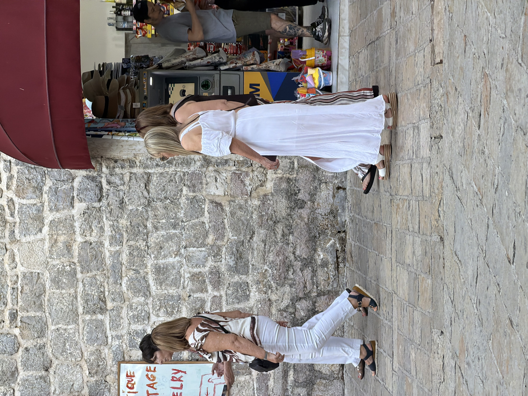 In Budva, Montenegro, a group of four people walks along a cobbled street in front of a stone wall and a store with a red awning. Two women in casual summer clothing walk from left to right. The woman on the far left wears white pants and a patterned blouse, carrying a black bag. The woman next to her wears a white off-the-shoulder dress and white sandals. Behind them, two more people are partially visible. One is a woman wearing a striped dress, and another is a man wearing a grey t-shirt and shorts, with tattoos on his legs. The store appears to sell hats, souvenirs, and other merchandise. The scene depicts a typical summer day in a tourist area.