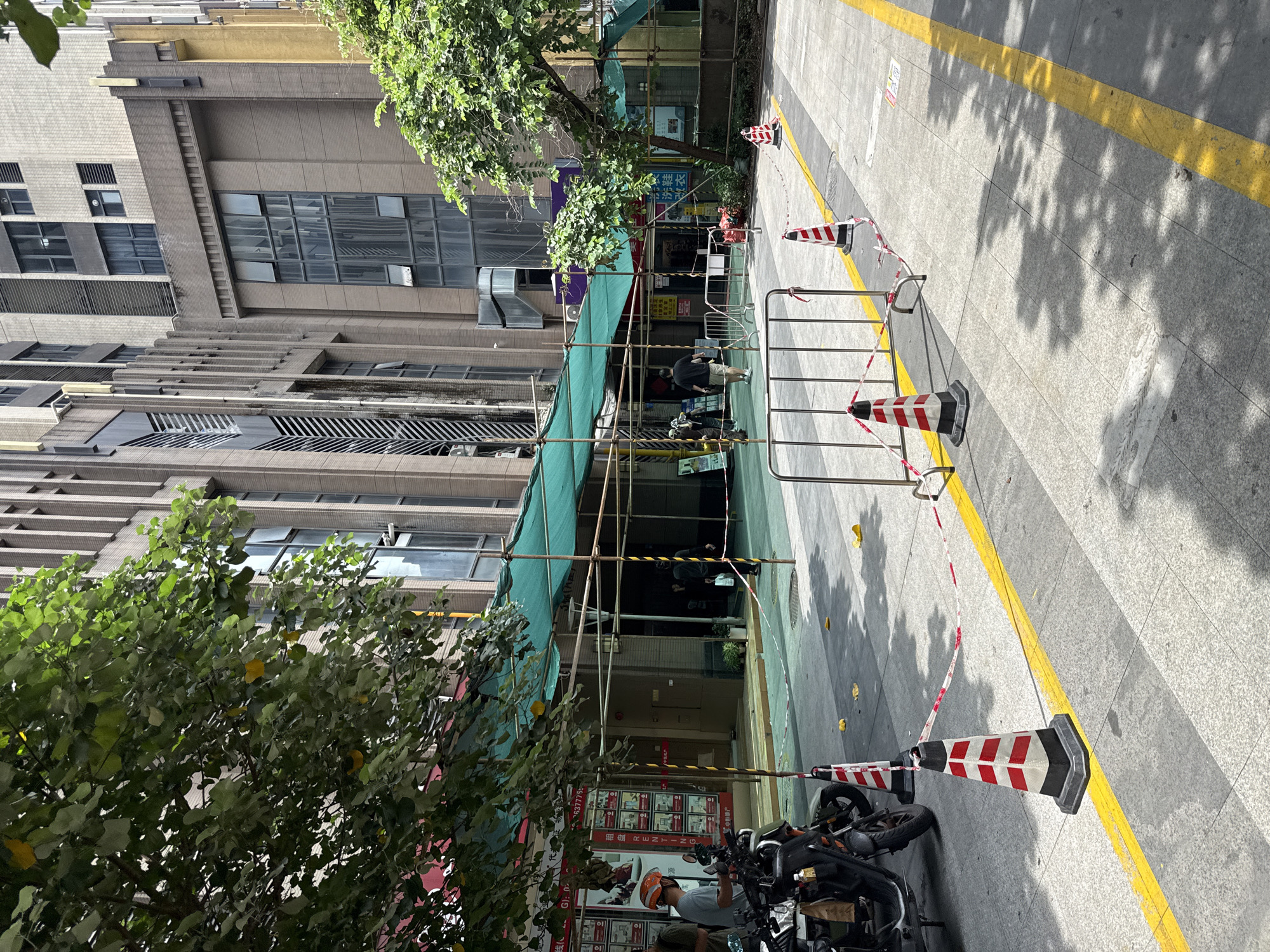 A street scene in Haizhu District, China, features a sidewalk with a yellow line and a series of construction barriers, traffic cones, and red and white caution tape. In the background, a tall, modern building with many windows dominates the cityscape. To the left, a tree with green leaves partially obscures the view. In the foreground, a motorcycle and a person wearing an orange helmet are visible. A green canopy, supported by scaffolding, extends over a section of the street, suggesting ongoing construction or maintenance. Several individuals are seen walking or standing under the canopy. The ground is paved with tiles, and scattered yellow leaves are present.