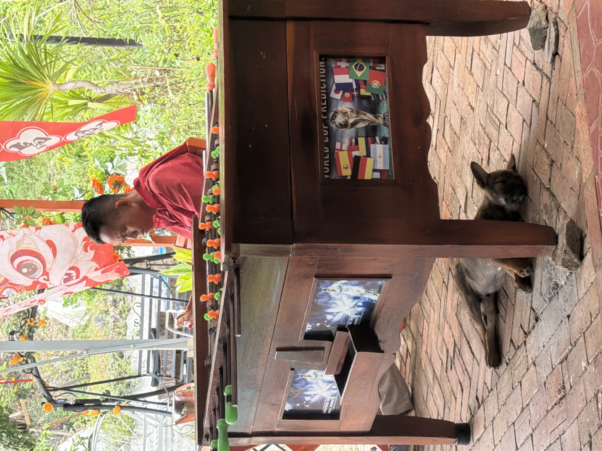 A man in a red shirt is playing foosball on an outdoor patio. The foosball table has a "World Cup Prediction" graphic on its side, along with soccer-themed decorations. A small dog is sleeping near the table's legs on a brick patio. The environment includes greenery, flags with decorative patterns, and a glasshouse-like structure in the background. It is unclear that the location is specifically in the city of Đà Nẵng, Vietnam.