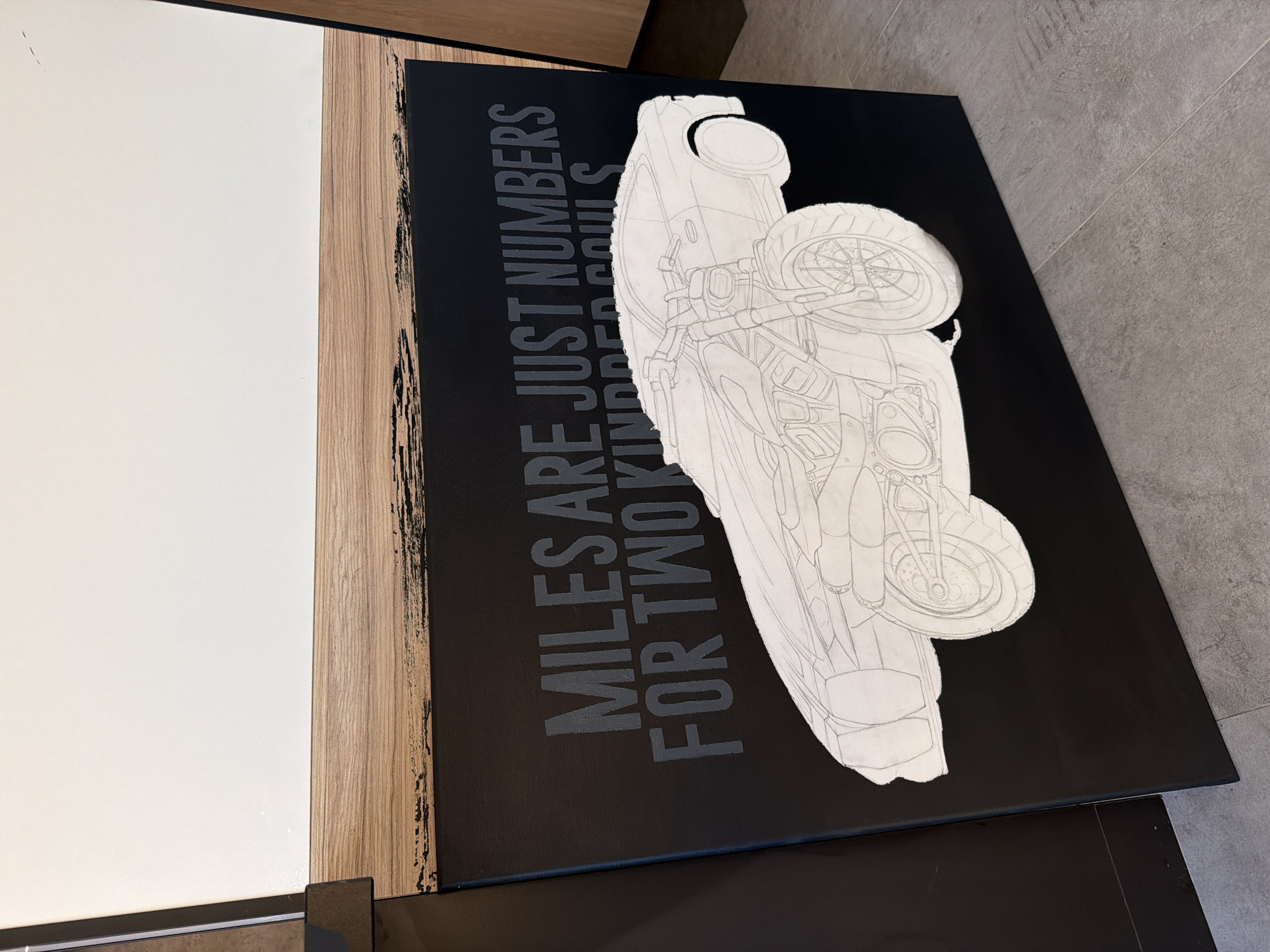A black canvas featuring a gray drawing of a motorcycle and the text "Miles are just numbers for two kinder souls" is positioned atop a dark surface. The canvas is positioned adjacent to a white surface, separated by a wood-grained material. The photograph is taken indoors.