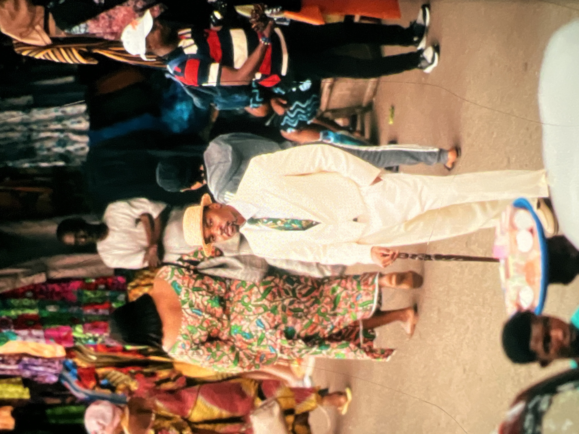 The image captures a vibrant street scene, likely in a bustling market in Lagos, Nigeria. In the foreground, a man dressed in a cream-colored suit and a straw hat stands with a cane in his right hand, looking towards the camera. He wears a green and white patterned tie. To his left, a woman in a colorful, patterned dress walks away from the camera. Behind her, another woman in a pink and yellow patterned outfit is partially visible. Further in the background, several other individuals are present, including a man in a white shirt and another wearing a striped polo shirt and a white baseball cap. Stalls displaying an array of colorful fabrics are visible behind the people. The ground appears to be paved with a rough surface, typical of a street or market walkway. The overall impression is one of lively commerce and daily life in an urban Nigerian setting.