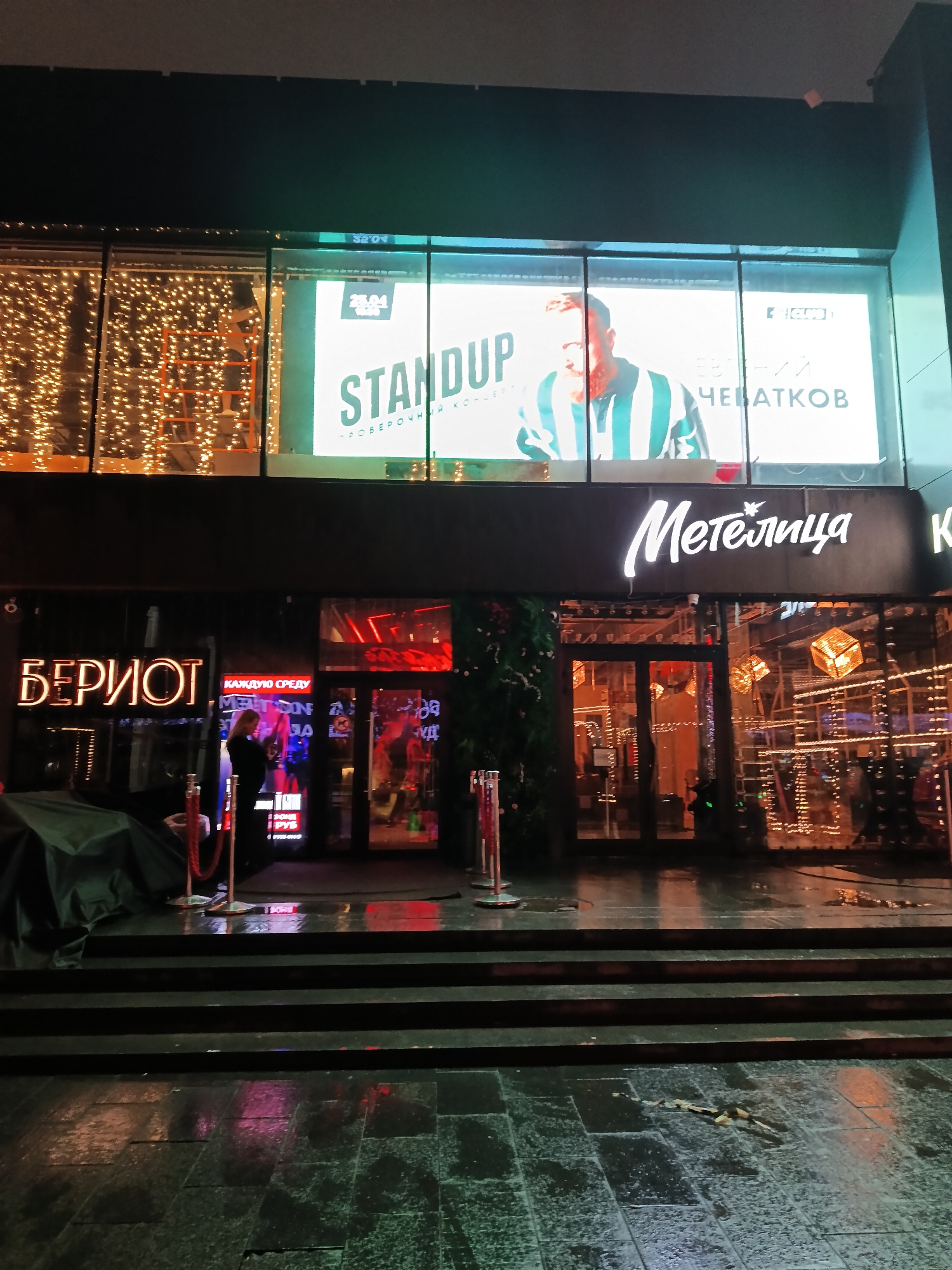 The image captures a nighttime scene in Moscow, Russia, featuring the entrances to two establishments. On the left, the signage reads "БЕРІОТ" in illuminated lettering. A person is standing near the entrance, in front of a red rope barrier. To the right is the entrance to "Метелица" with the name prominently displayed in a cursive font. Above the entrances, a large billboard advertises a "STANDUP" performance by Евгений Чебатков (Evgeniy Chebatkov). Decorative lights are visible in the windows above and inside both establishments. The ground consists of steps and a wet, reflective pavement.