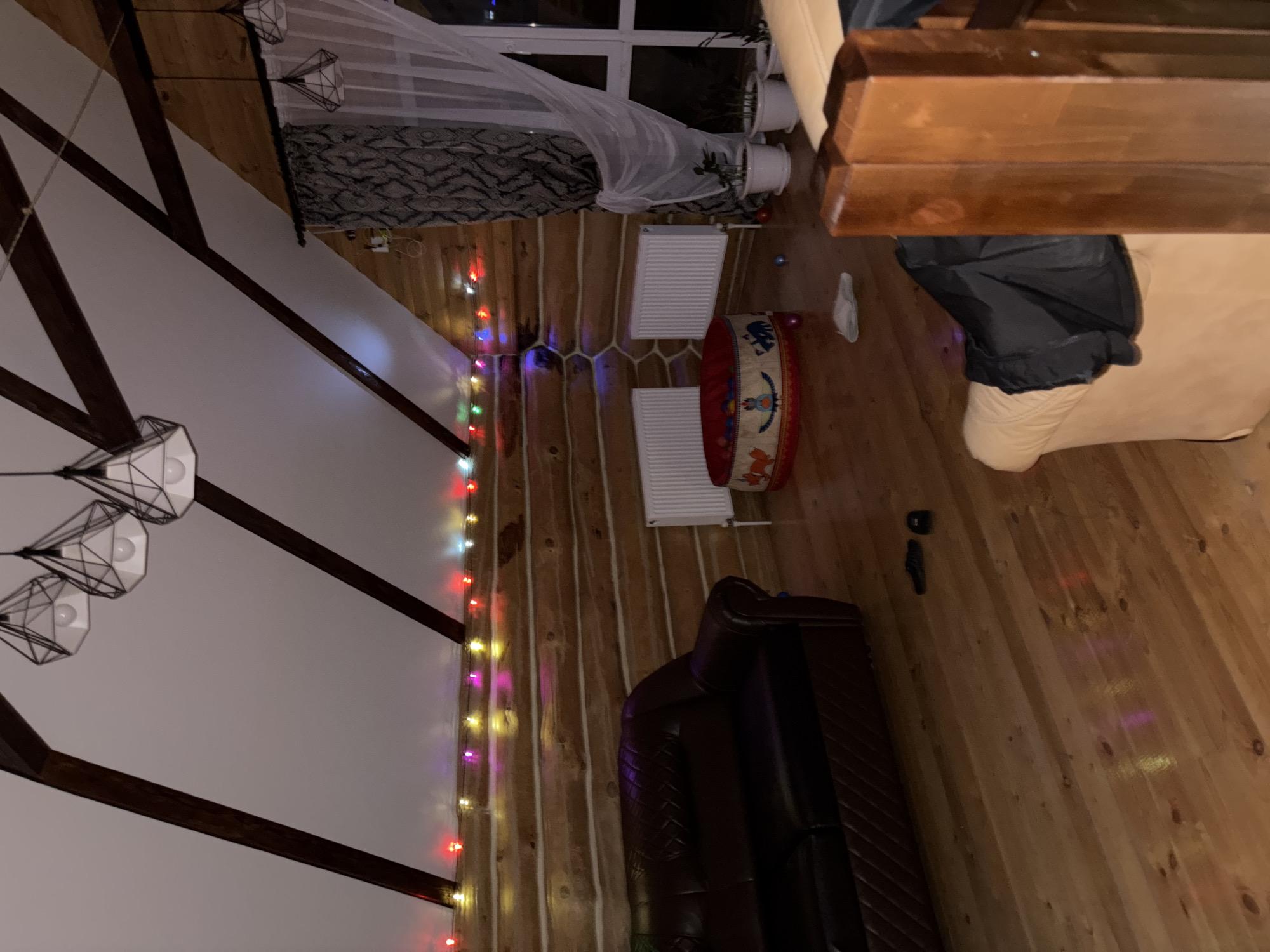 The image shows an interior space with wooden walls and floors, likely a house or cabin. The room has a pitched roof with dark wooden beams, under which hang modern geometric light fixtures. A string of colorful Christmas lights runs along the wall. There is a leather couch, a child’s drum, and two white radiators. The window is visible with translucent curtains and plants on the windowsill. Given the mention of Chaikovsky, Russia, it is possible that this room is located there.