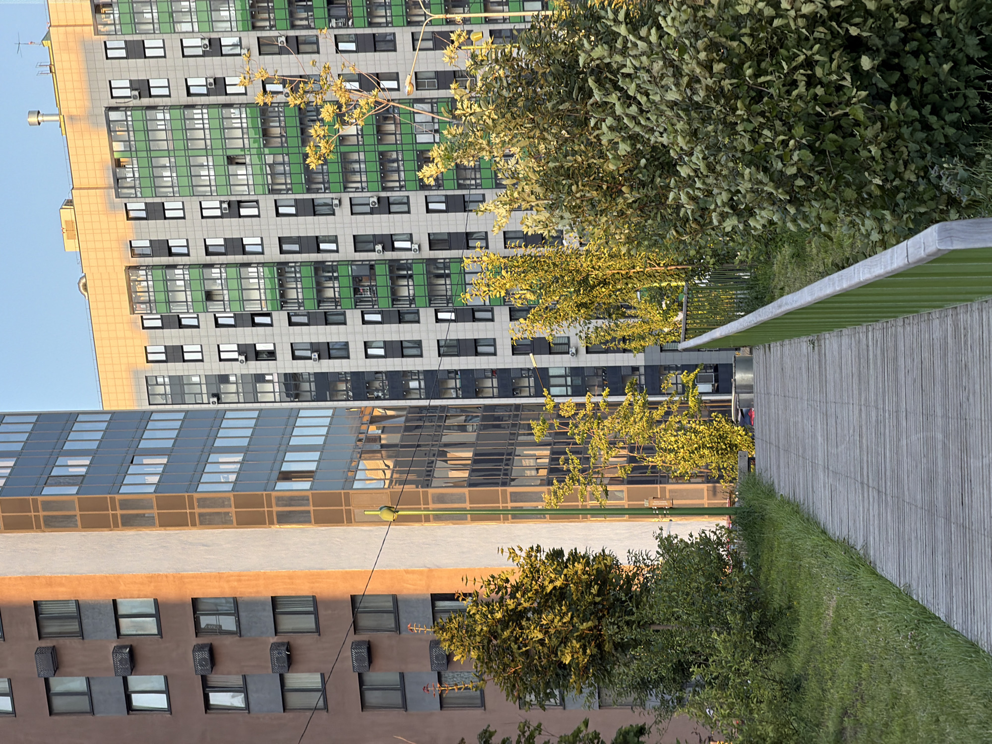 In an urban setting, a wooden pathway slopes upwards through a grassy area. On the left, a multi-story building with a brown facade is visible, punctuated by numerous windows and air conditioning units. Adjacent to it, another building features a cream-colored section and a glass facade with vertical brown elements. To the right, a contemporary apartment building with a yellow-orange top section stands, its facade characterized by grey panels, white window frames, and balconies with green railings. Lush trees with green and yellow foliage line the pathway, with their branches reaching towards the buildings. A light green railing follows the curve of the pathway. The sky is clear and blue.