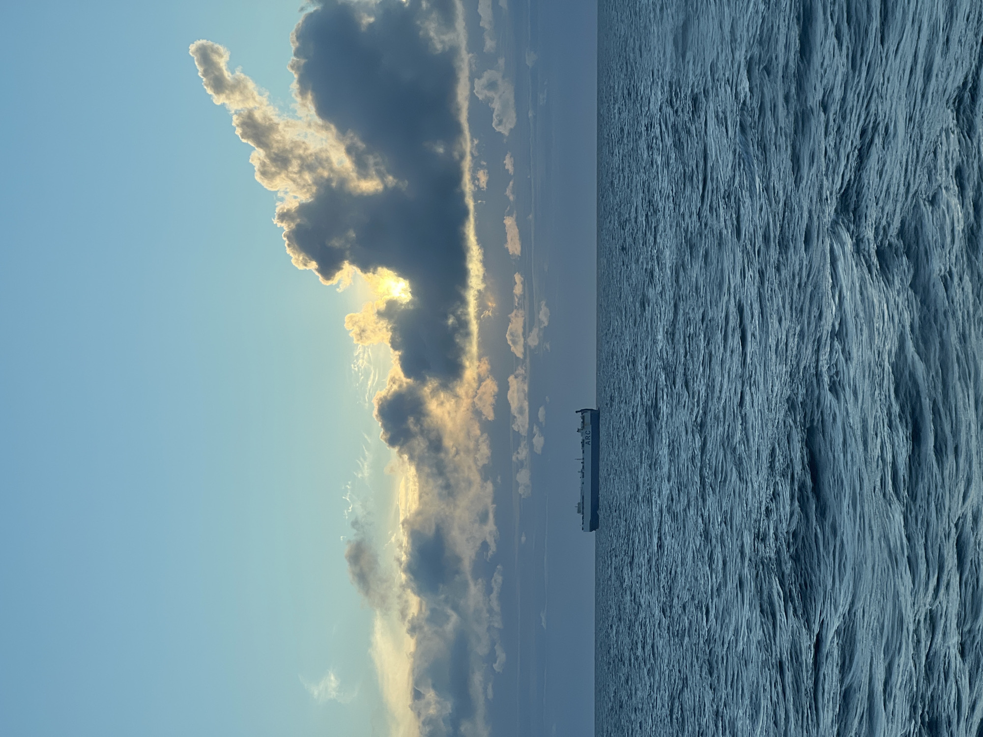 A car carrier ship, identified by the name "ARC" visible on its hull, sails on the deep blue, choppy sea under a partially cloudy sky. The sun is visible through a gap in the clouds, casting a warm glow on the scene. The horizon is clearly defined, separating the ocean from the sky. The ship is the only discernible object in the expansive seascape. The environment is a maritime setting, possibly offshore. The location is an unknown area of Poland.