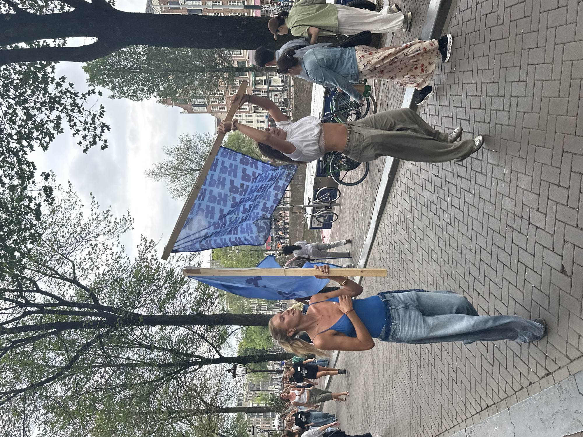A group of people are walking along a canal in Amsterdam, Netherlands. The people are carrying flags and appear to be participating in a demonstration or parade. The canal is lined with buildings, and there are trees along the street. The weather is cloudy. A woman in the foreground is wearing a blue bodysuit and jeans and holding a blue flag on a wooden pole. Another woman behind her is wearing a white crop top and loose-fitting pants and holding a larger blue flag. Several other people are visible in the background, some of whom are also carrying flags. Bicycles are parked along the canal.