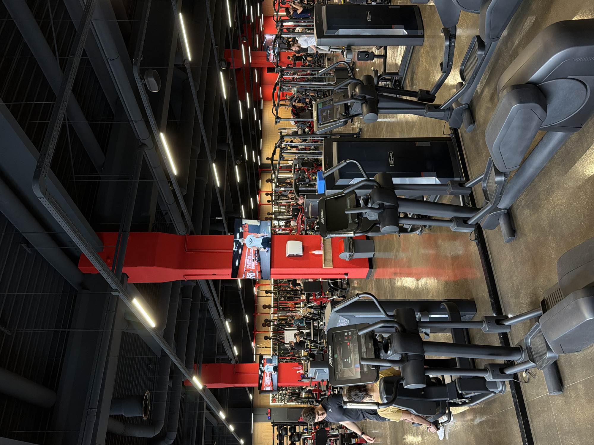 The image depicts the interior of a gym in Plovdiv, Bulgaria. Rows of exercise machines, including elliptical trainers and weight machines, are arranged in parallel lines. Several individuals are using the equipment. The gym features a high ceiling with exposed black infrastructure, including pipes and beams, punctuated by linear fluorescent lighting. Red ductwork is also visible. The floor appears to be a textured, composite material.