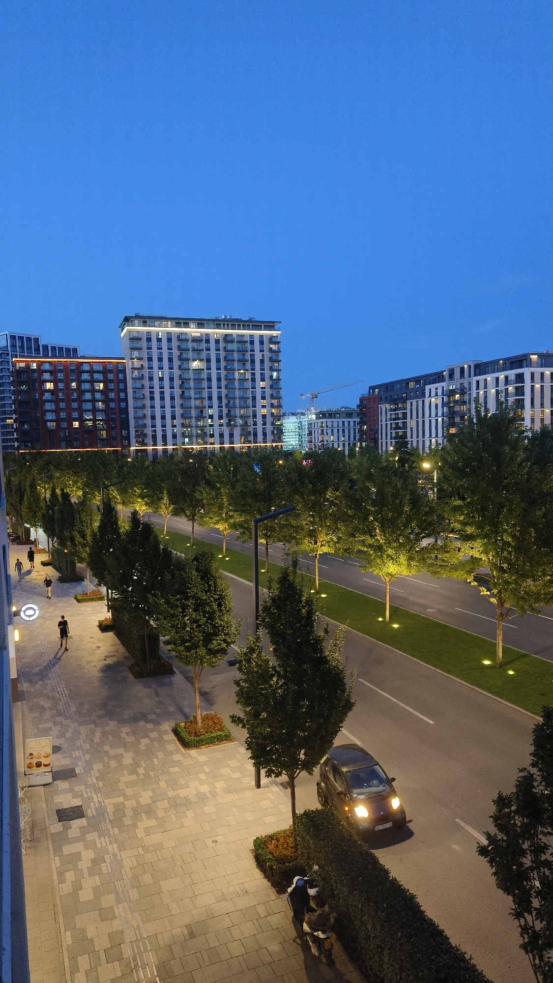 This is a vertical, high-angle, daytime photograph of a modern urban street scene in Belgrade, Serbia. The sky is a deep blue, indicating late evening or early morning. In the background, several multi-story residential buildings with balconies and illuminated windows are visible. One building, towards the left, is red brick with orange lighting accents, while others are lighter-colored with white balconies. A construction crane can be seen behind some of the buildings.
The foreground features a wide pedestrian walkway paved with light gray rectangular stones. Several trees are planted along the edge of the walkway, interspersed with small, neatly manicured hedges and planters. Streetlights, mounted on tall poles, illuminate the walkway, and small ground lights are embedded in the grass beside the road.
A paved road runs parallel to the walkway, bordered by a green lawn. A dark-colored compact car with its headlights on is driving on the road, heading away from the viewer. On the walkway, two individuals are walking away from the camera, and another person is walking towards the camera. A scooter with two individuals on it is parked along the hedge on the right side of the walkway. A small display with images of pastries is positioned on the left side of the walkway. The overall impression is of a well-maintained, contemporary urban environment.