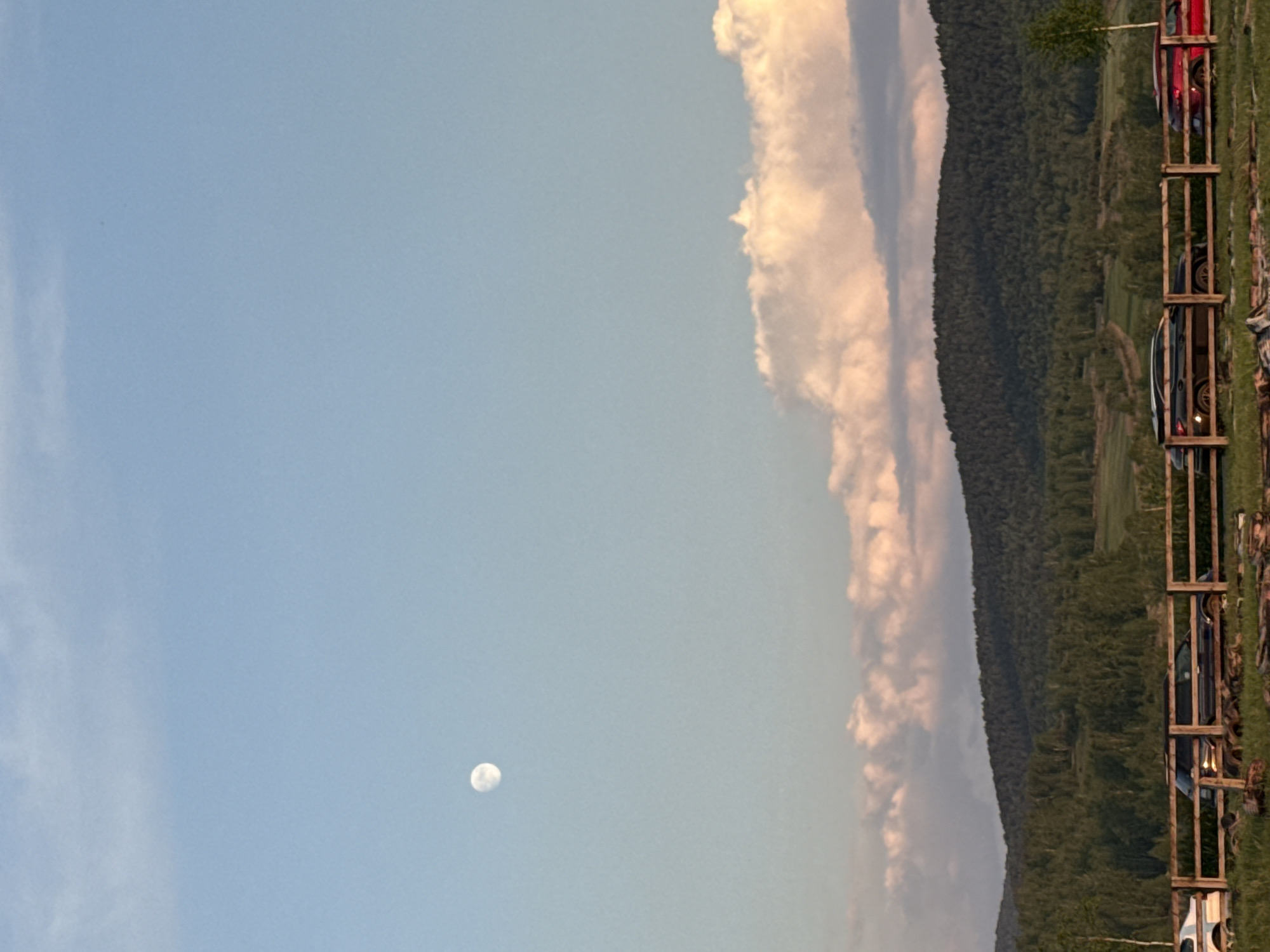 The image shows a landscape in Кузнецово, Russia. The sky is predominantly blue with clouds on the right side of the image. The moon is visible in the lower left quadrant. Adjacent to the clouds is a densely forested hill. On the right, a fence is visible, with cars parked along it.