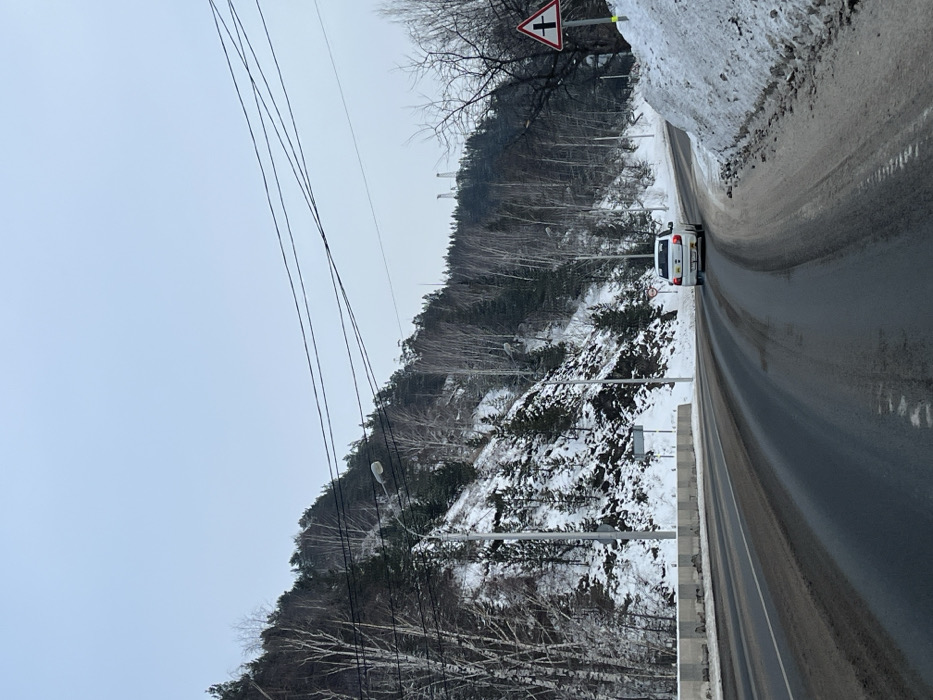 The image depicts a winter scene on a paved road in Слизнево, Russia, under an overcast, light grey-blue sky. The road, appearing damp and soiled, features at least two lanes and curves slightly to the right as it recedes into the distance. A white concrete barrier runs along the left edge of the road, separating it from a steep, snow-covered hillside. This hillside is densely vegetated with a mix of dark evergreen trees and bare deciduous trees, primarily birches.
A single white sedan, possibly a Renault Logan, is traveling away from the viewer in the rightmost lane. Several grey streetlights with oval luminaires are spaced along the left side of the road, following its curve. Multiple dark power lines traverse the upper portion of the frame, spanning above the road and hillside. In the far distance on the horizon, two distinct industrial structures resembling transmission towers or pylons are visible atop the tree line.
On the right shoulder of the road, a significant accumulation of snow and ice is present. A triangular warning sign with a red border and a black symbol indicating a side road joining from the right is prominently displayed on the right. A smaller, circular sign with a red border and a white background, featuring an indistinct black symbol, is visible on the left side near the white vehicle. The overall environment suggests cold temperatures with snow present on the ground and trees. No individuals are visible in the frame.