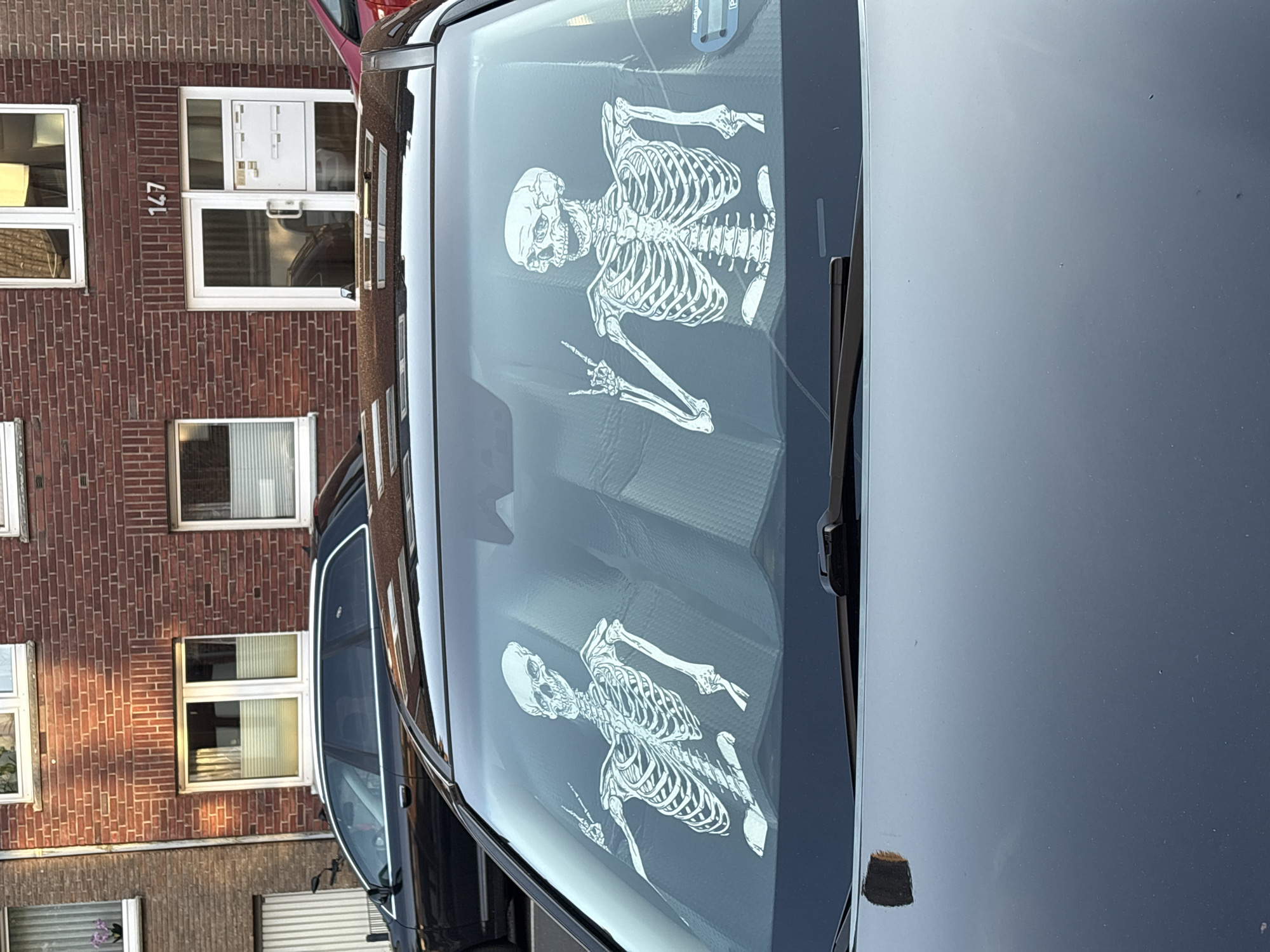 The image shows the windshield of a dark car with a reflective sun shade inside, on which two white skeleton figures are printed. The skeletons are depicted in a casual pose, with one hand making a "peace" or "victory" sign. Outside the car, a brick building with the number "147" is visible. Based on the architectural style and the presence of German license plates on vehicles that may be present, the location is determined to be Mönchengladbach, Germany.