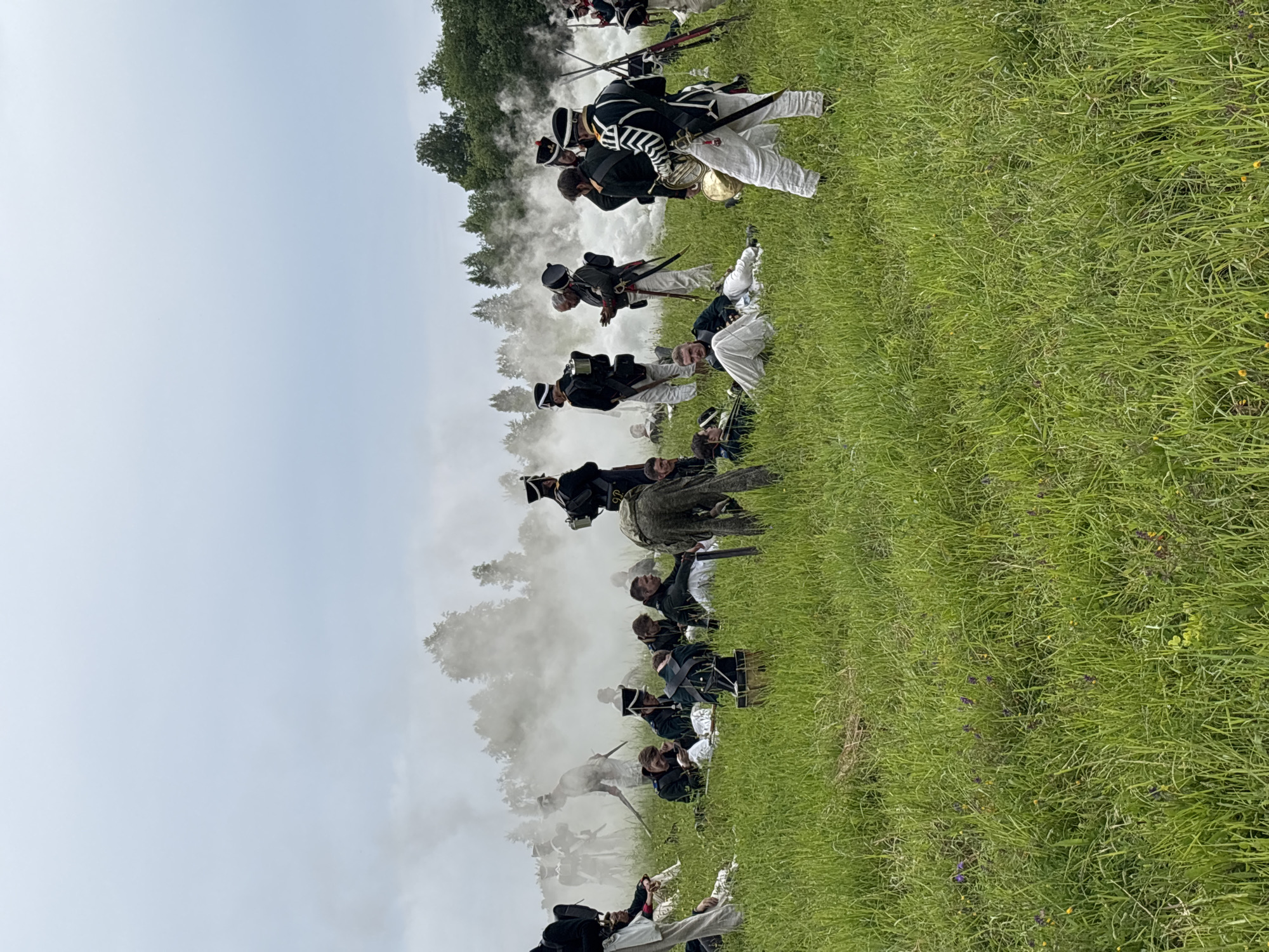 The image depicts a reenactment of a historical battle, possibly from the Napoleonic era, taking place in a grassy field, likely in Goncharovo, Russia. A group of individuals dressed in period military uniforms are scattered across the field. Some are standing, while others are seated or lying down, suggesting a pause in the action or fallen soldiers. Smoke billows in the background, adding to the atmosphere of a battlefield. The participants are wearing dark jackets, white trousers, and distinctive hats. Some are holding weapons, and one person appears to be playing a musical instrument, possibly a horn. The environment is a natural landscape with green grass and trees in the distance under an overcast sky.