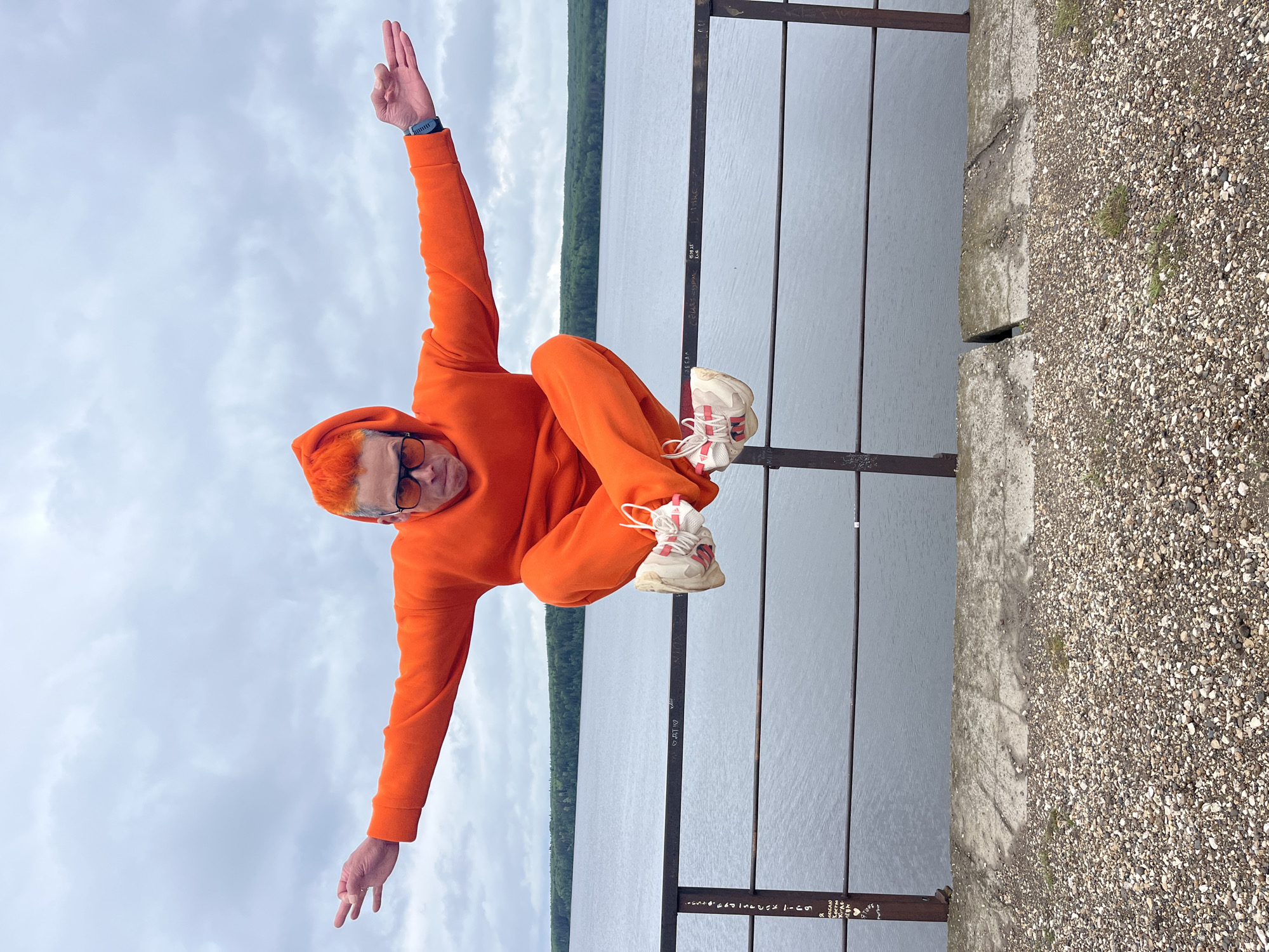 A person in an orange hooded jumpsuit and white sneakers is perched on a metal railing along a concrete embankment. The person has short, dyed orange hair and is wearing glasses. Their right arm is raised upward, and their left arm extends downward. In the background is a body of water with a tree-lined shoreline and a cloudy sky. The embankment consists of concrete sections and gravel. The location is Chaikovsky, Russia.